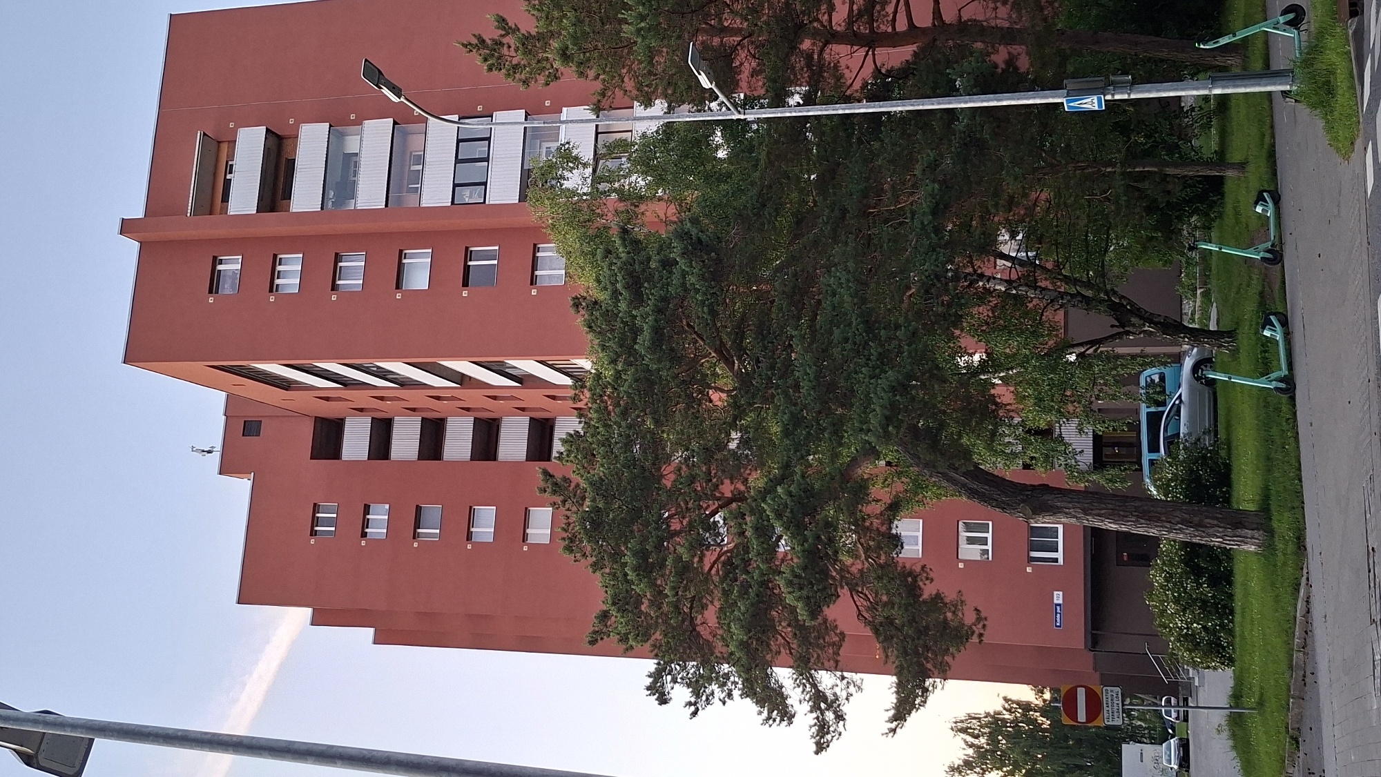 In the city of Tallinn, Estonia, a tall, reddish-orange apartment building dominates the scene. The building features multiple floors with numerous windows and balconies. In front of the building, a variety of green electric scooters are parked on the sidewalk. A silver car and a light blue van are visible parked in front of the building. A traffic sign, a street lamp, and trees are also present in the environment. The sky is clear with a faint cloud.