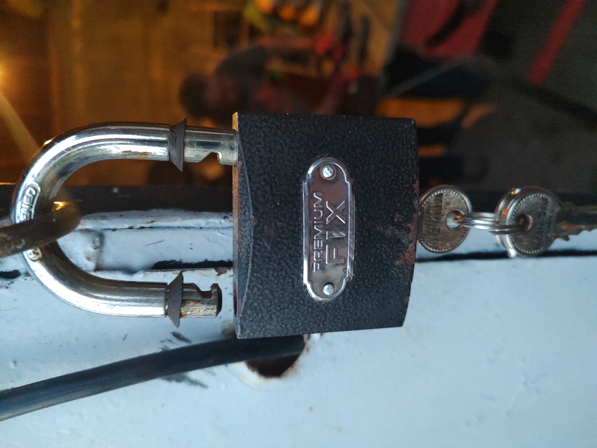 A close-up, eye-level shot captures a weathered, black, rectangular padlock with a silver metal plate on its front. The plate is embossed with the words "PREMIUM FIX" in an arch above the word "FIX" in larger letters. The padlock's silver shackle is partially open, revealing its locking mechanism. Two keys hang from a metal ring attached to the bottom of the padlock. The padlock is secured to a light blue metal surface, likely a door or gate, which shows signs of rust around a circular hole near the shackle. To the left of the padlock, a dark gray, cylindrical rod runs vertically. The background is blurred and appears to be an indoor or semi-outdoor setting with warm lighting and indistinct figures of people, suggesting a workshop or storage area. The overall impression is one of an outdoor, possibly neglected, urban environment in Kochmar, Bulgaria.