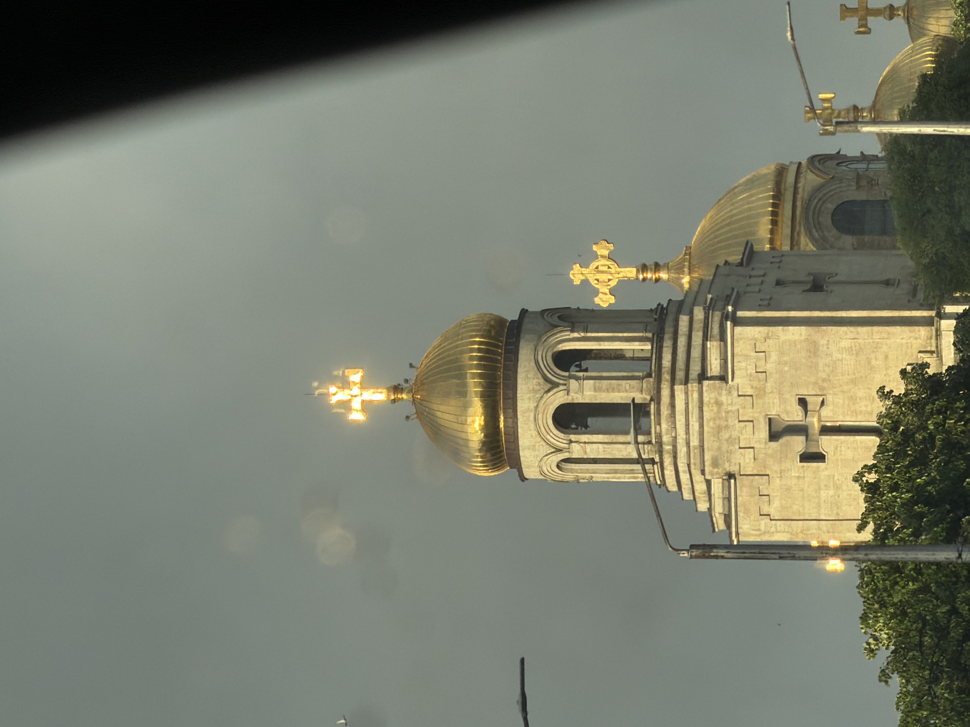 A grand architectural structure, likely an Eastern Orthodox church or cathedral, is prominently featured in this image, viewed from a slightly upward angle. The building is characterized by its multiple gleaming golden domes, each topped with an ornate golden cross.
The most prominent dome in the foreground is intricately fluted, reflecting a warm, golden sunlight that suggests either late afternoon or early morning. This golden light brightly illuminates the crosses and domes, contrasting with the muted, soft gray-blue sky. The building's walls are constructed from a light-colored stone or concrete, adorned with elaborate architectural details, including arched openings in what appears to be a bell tower section, and decorative elements on the facade. A distinct cross shape is also integrated into the stonework of the building itself, visible below the domes.
Further back and to the right, another large golden dome and cross are partially visible, peeking out from behind green foliage. Trees are present on the right side of the frame, partially obscuring parts of the building and adding a touch of natural color. Elements of urban infrastructure, such as a utility pole or wires, are also discernible in the lower and middle sections of the image, suggesting an urban or suburban setting. No people or specific activities are depicted, and no text is visible. The overall mood is one of serenity and architectural majesty.