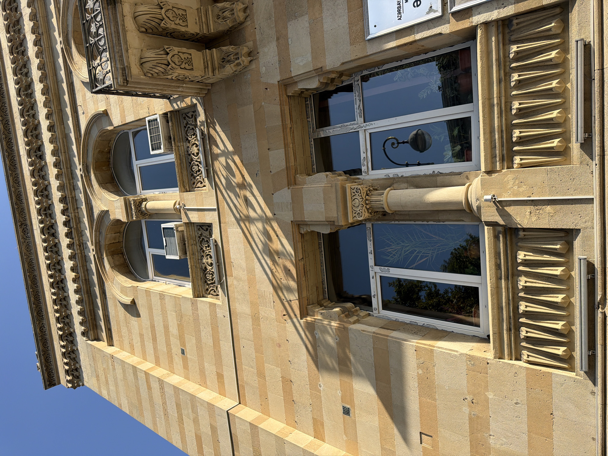 The image shows a low-angle, full-frame view of the facade of a building, likely in Sabail Raion, Azerbaijan, under a clear blue sky. The building is constructed of light-colored stone with horizontal bands of varying shades, creating a striped effect. The architecture features ornate detailing, including arched windows, decorative moldings above the windows, and elaborate carved columns supporting the upper levels.
In the upper section of the building, two arched windows are visible, each fitted with a white window frame. An air conditioning unit is installed in the lower part of the right window. A portion of a wrought-iron balcony with intricate scrolling patterns extends from the right side of the building.
Below the upper windows, a large, rectangular window dominates the center of the frame. This window has a white frame and is divided into multiple panes, reflecting the sky and trees outside, as well as an interior view of potted plants and a decorative lamp. Another similar window is partially visible to the left of this main window.
A decorative stone element, resembling a series of pointed projections, runs horizontally below both the upper and lower window levels. The building's facade is casting long shadows from an unseen source, suggesting it is late afternoon or early morning. A small, dark surveillance camera is mounted on the wall to the left of the main window. On the far right, a portion of a sign with text, including "AZƏRBAYCAN" and other characters, is visible, further indicating its location in Azerbaijan. The overall impression is one of an older, well-maintained building with classical architectural influences.