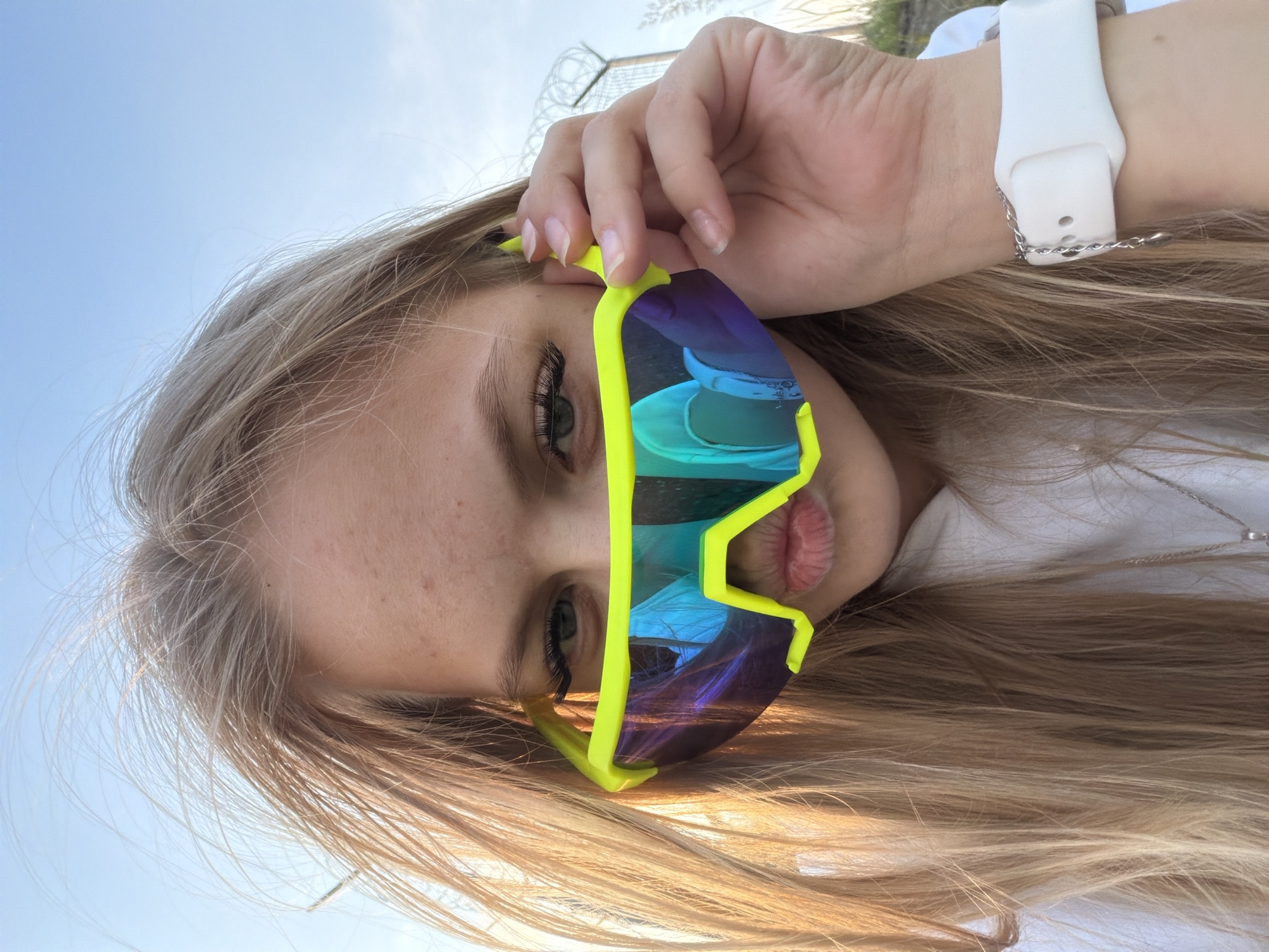 The image shows a close-up selfie of a young woman wearing bright neon yellow sport sunglasses with blue and purple mirrored lenses. Her hair is long and blonde, and it is blowing in the wind. She is wearing a white hoodie and a silver necklace. On her left wrist, she wears a white smartwatch with a small silver charm hanging from the strap. The background is a bright blue sky with some wispy clouds. In the upper right corner of the image, a barbed wire fence is visible. The scene appears to be outdoors, possibly in a park or open area, on the outskirts of Chaikovsky, Russia, given the presence of the fence which could indicate an industrial or restricted area. The lighting suggests it is daytime, possibly late afternoon due to the warm glow on her hair.