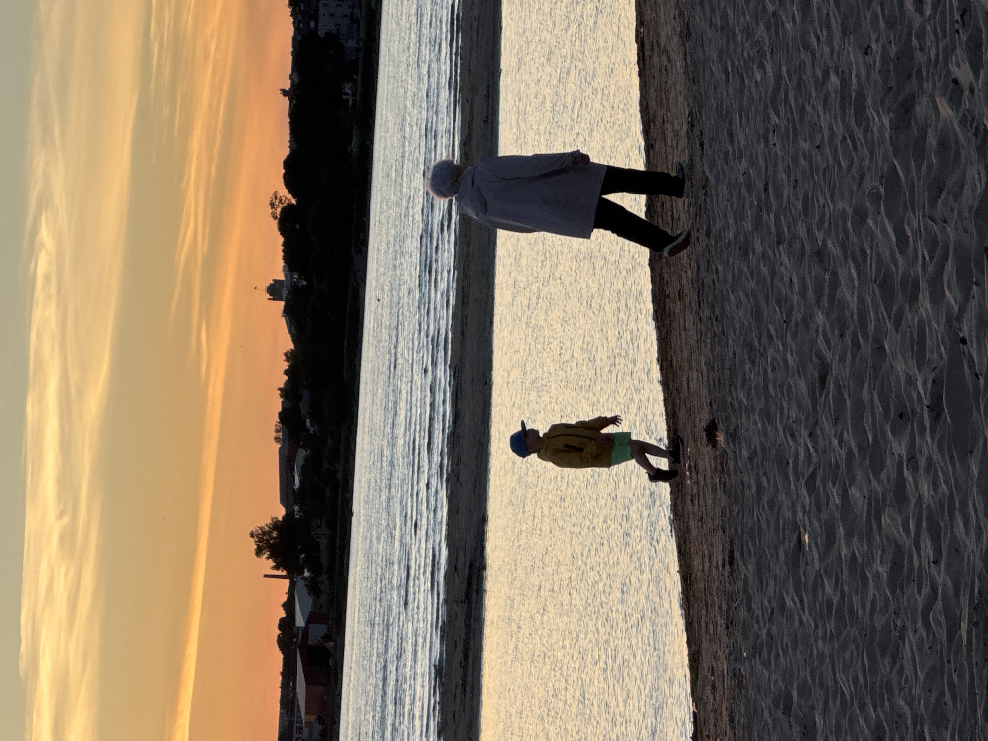The image shows two people, a child and an adult, walking on a sandy beach alongside a body of water at sunset. The sky is a vibrant orange and yellow, with wispy clouds. In the background, across the water, there is a silhouetted shoreline with trees and buildings, including what appears to be a tall tower with a flag. The scene is likely Tallinn, Estonia, given the characteristic waterfront architecture and sunset hues common to the Baltic Sea region. The child, wearing a yellow jacket and blue cap, is walking ahead of the adult, who is wearing a grey hooded jacket and dark pants. The water is calm, reflecting the colors of the sky. The sand is textured with footprints and ripples.