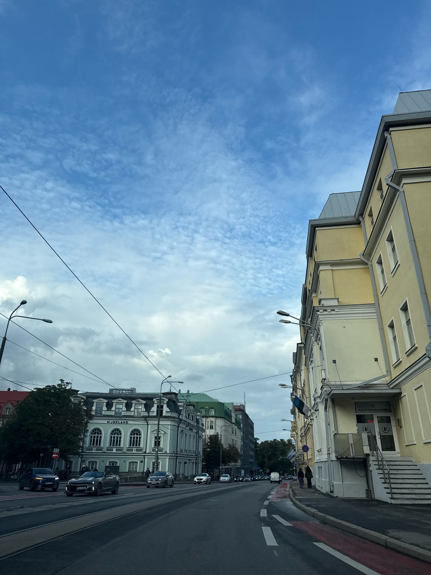 The image depicts a street scene in Tallinn, Estonia, likely in the city center. On the right side of the street, a prominent light yellow building with white decorative elements and multiple windows is visible. This building features a side entrance with stairs leading up to a doorway marked with the "P" logo. Flags, including the Estonian flag, are displayed on the building's facade. Across the street, on the left, a light green building with a grey roof, dormer windows, and the word "WORKLAND" inscribed above its arched windows stands out. Further down the street, other buildings with varied architectural styles and colored roofs are visible, interspersed with trees. The street itself is paved with asphalt and has tram tracks. Several cars, including a dark sedan, a silver convertible, and other vehicles, are driving or parked on the street. People are seen walking on the sidewalk near the entrance of the yellow building and further down the street. The sky above is partly cloudy with scattered white clouds against a blue background. Overhead utility lines are also visible crisscrossing the frame.