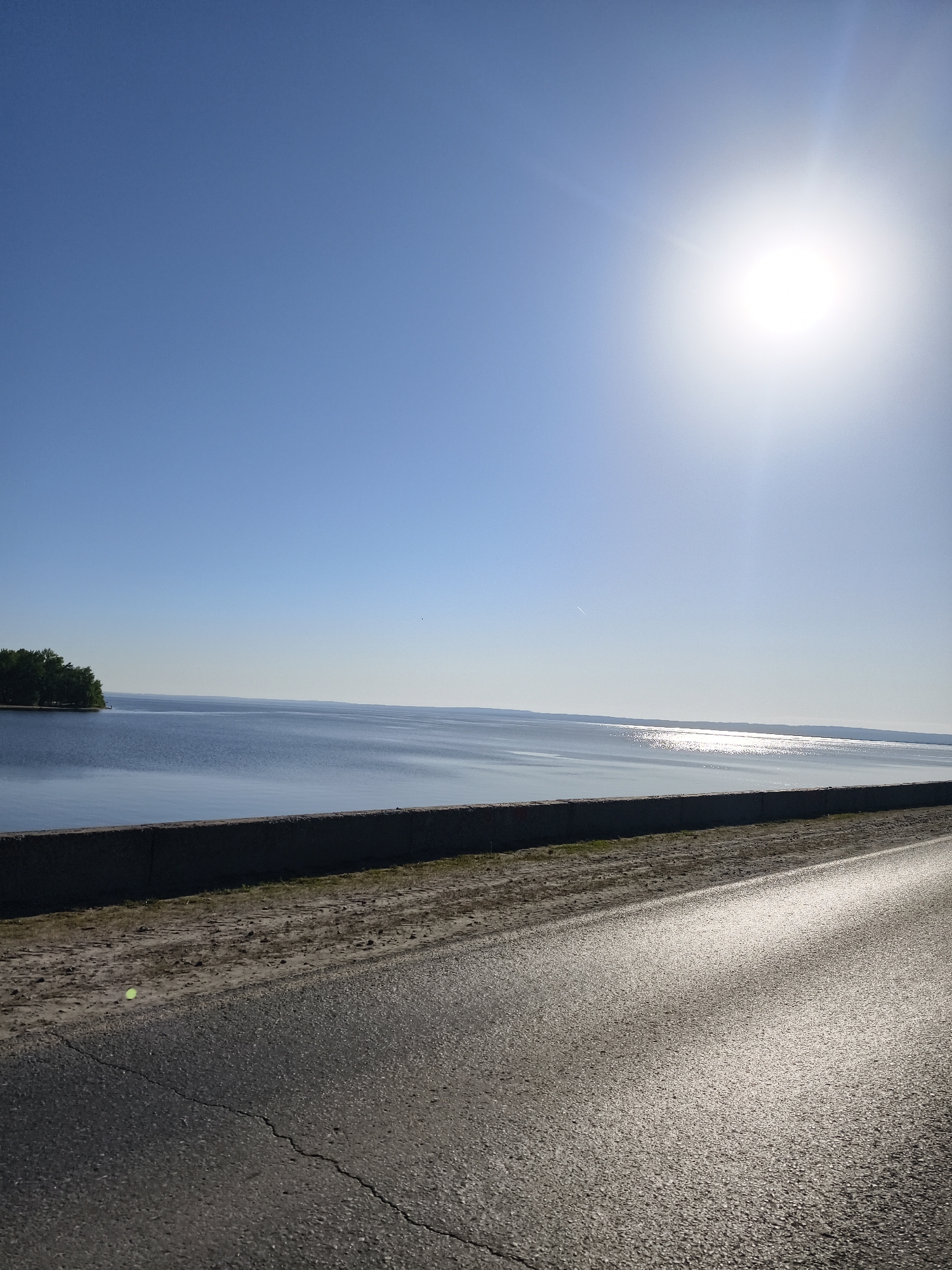 The image shows a view of the Volga River in the city of Noviy, Russia. The sky is clear and blue with a bright sun. The river is calm, reflecting the sunlight. A concrete barrier separates the river from a paved road in the foreground. A small patch of green trees is visible on the left side of the river. The road is gray and shows some cracking.