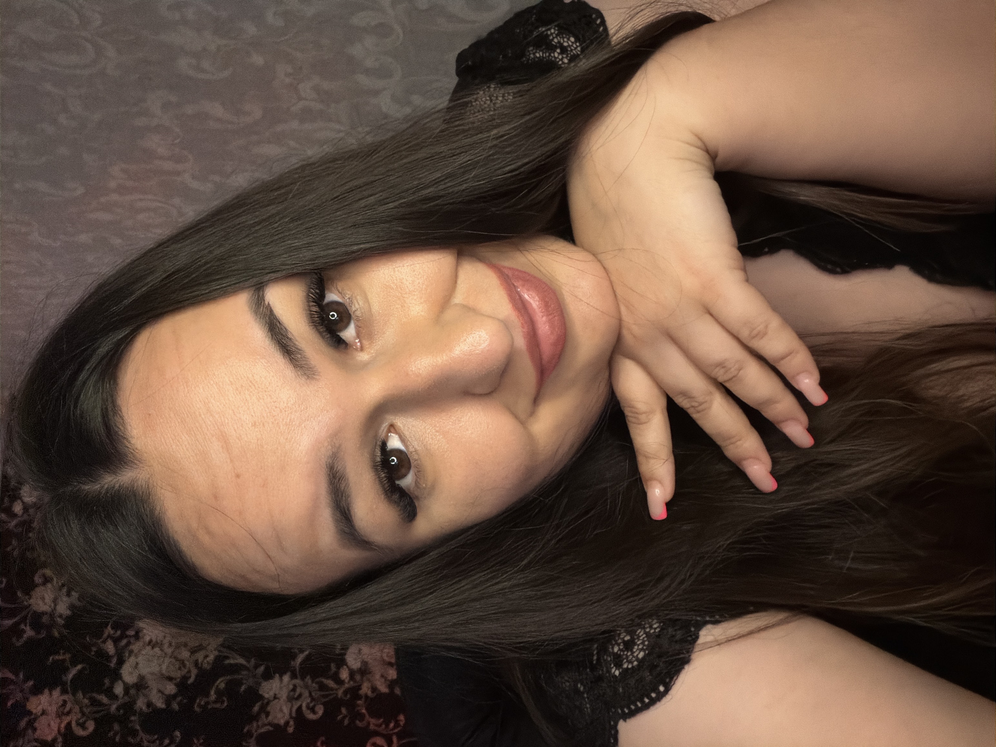 A close-up, self-portrait shows a woman with long, dark brown hair, resting her chin on her hand. She is wearing a black lace top. Her eyes are dark brown, and she has long, dark eyelashes. Her lips are painted a glossy pink. Her nails are light pink with a French tip. The background is a patterned wallpaper with darker floral motifs on the left and a lighter textured pattern on the right. The lighting is warm and soft, casting a gentle glow on her face.