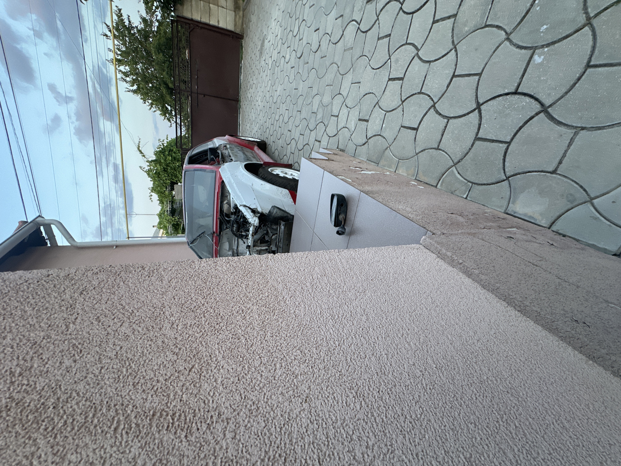 An incomplete, red and white car sits on a paved driveway in Comrat, Moldova. The car's hood is open, revealing the engine compartment. The driveway is made of grey, interlocking pavers arranged in a wavy pattern. A textured, beige-colored wall is on the left side of the image, partially obscuring the car. In the background, a dark brown metal gate stands closed. Green trees and a cloudy sky are visible behind the gate. A white gutter runs along the top edge of a building on the left. A black car part rests on the edge of a small step in front of the beige wall.