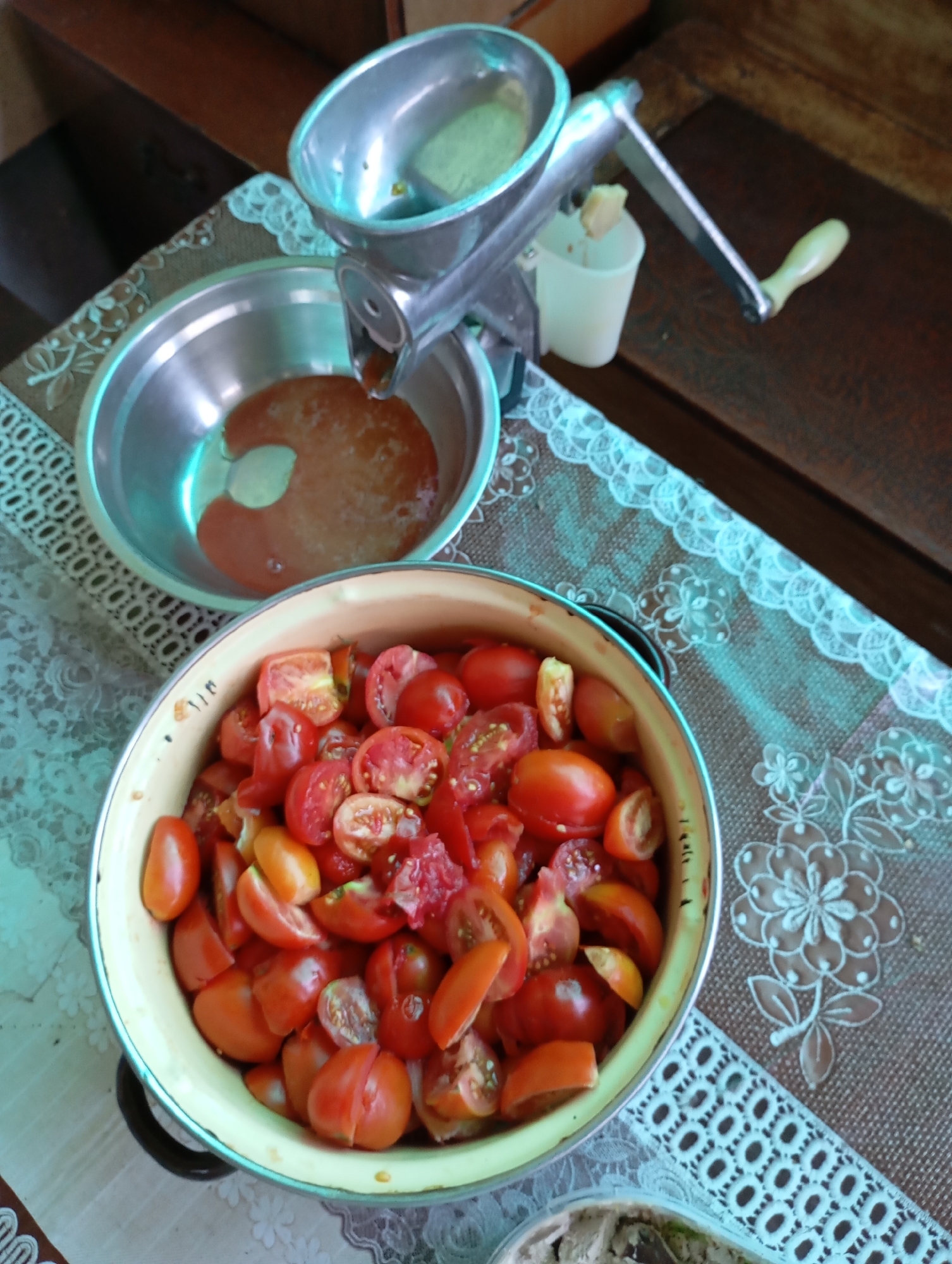 In Horishni Plavni, Ukraine, a still-life image depicts a kitchen scene focused on tomato preparation. A manual tomato press, made of aluminum with a wooden crank handle, is positioned on a wooden surface. Below the press, a metal bowl contains some reddish liquid, presumably tomato juice or pulp. In the foreground, a large, light-colored pot overflows with chopped tomatoes. The table is adorned with a brown tablecloth featuring a white lace trim and floral pattern. The overall impression is one of domestic food preservation.