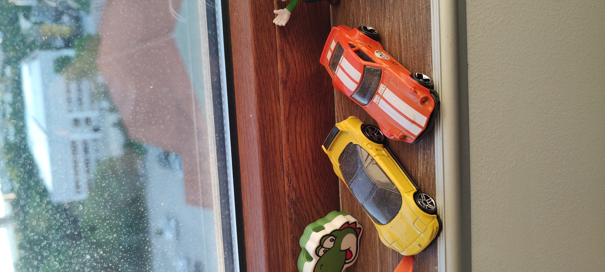 The image depicts a window sill with various objects placed on it. An orange toy car with white stripes and a "24" on the hood is visible, along with a yellow toy car. Additionally, there is a Yoshi figurine from the Mario franchise. The window has a view of buildings and greenery outside.