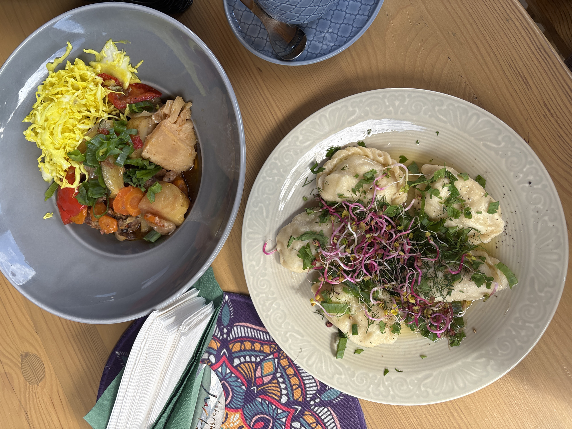 This is an overhead shot of two plates of food on a wooden table. The plate in the foreground is a light cream color with an embossed floral pattern around the rim. It contains several pierogi topped with chopped parsley and a pile of pink and green sprouts. The second plate, a larger gray bowl, is in the background and contains a stir-fry with what appears to be chicken, potatoes, carrots, red peppers, and green onions, topped with shredded yellow cabbage. To the left of the foreground plate, there are folded white napkins on top of a green napkin and a patterned fabric coaster. To the right of the gray bowl, a blue saucer holds a blue cup with a wooden spoon inside. The setting appears to be a restaurant or cafe in Górka, Poland.