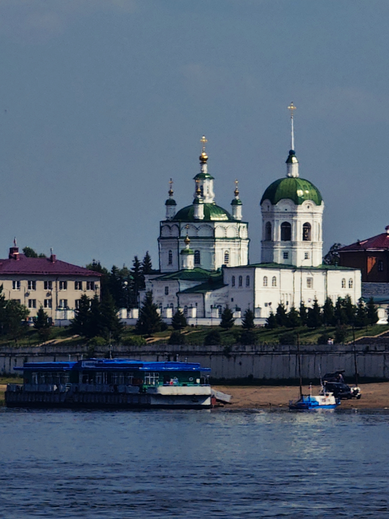 The image depicts a waterfront scene in the city of Yeniseysk, Russia. In the background, a large, white church with green domes and golden crosses stands prominently against a clear blue sky. Adjacent to the church is a building with a reddish-purple roof and multiple windows. In the foreground, a blue and white boat is anchored on a sandy shore, with a dark SUV parked nearby. Further to the left, a long, blue, multi-story boat or ferry is docked along the waterfront. The water in the foreground is a deep blue, reflecting the sky above. Trees are scattered throughout the landscape, dotting the hills behind the church and along the shoreline.
