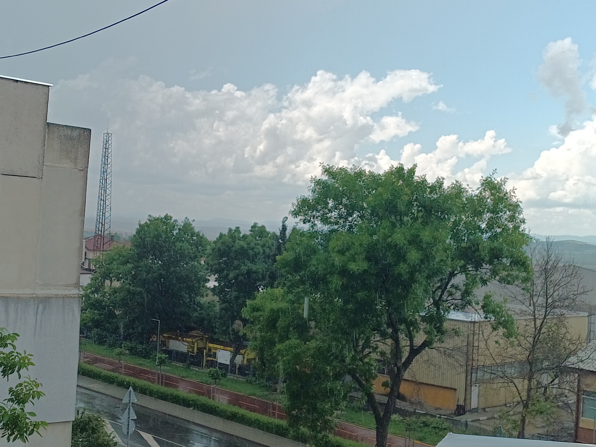 The image captures an outdoor, elevated perspective of an urban-industrial landscape in the city of Unknown, Unknown, under an overcast but bright sky with visible rainfall. The upper portion of the frame features a partly cloudy sky, with patches of light blue interspersed with numerous white and grey cumulus clouds. A thin, dark cable or wire traverses the top-left corner.
To the left, a segment of a light-colored, possibly concrete or stucco building, with some vertical streaking and discoloration, occupies the foreground. Behind this building, a tall, metal lattice communications tower with an antenna-like structure at its apex is prominently visible against the sky.
The mid-ground is dominated by lush green deciduous trees, which appear wet, with falling raindrops clearly visible, particularly around the large tree in the center-right. Below the trees, a wet, dark paved road or street is visible, featuring a white crosswalk marking and a triangular road sign. Adjacent to the road, a low reddish-brown barrier or fence runs parallel to what appears to be a track or path, possibly a railway line. Yellow heavy machinery or rail maintenance equipment is partially obscured by the foliage in this area.
In the background, beyond the immediate treeline, are several low-rise industrial buildings with corrugated metal siding, displaying shades of yellowish-beige and grey. A leafless tree stands in front of one of these structures. Distant, hazy hills or mountains are discernible on the horizon beneath the cloudy sky. No human figures are present in the scene.