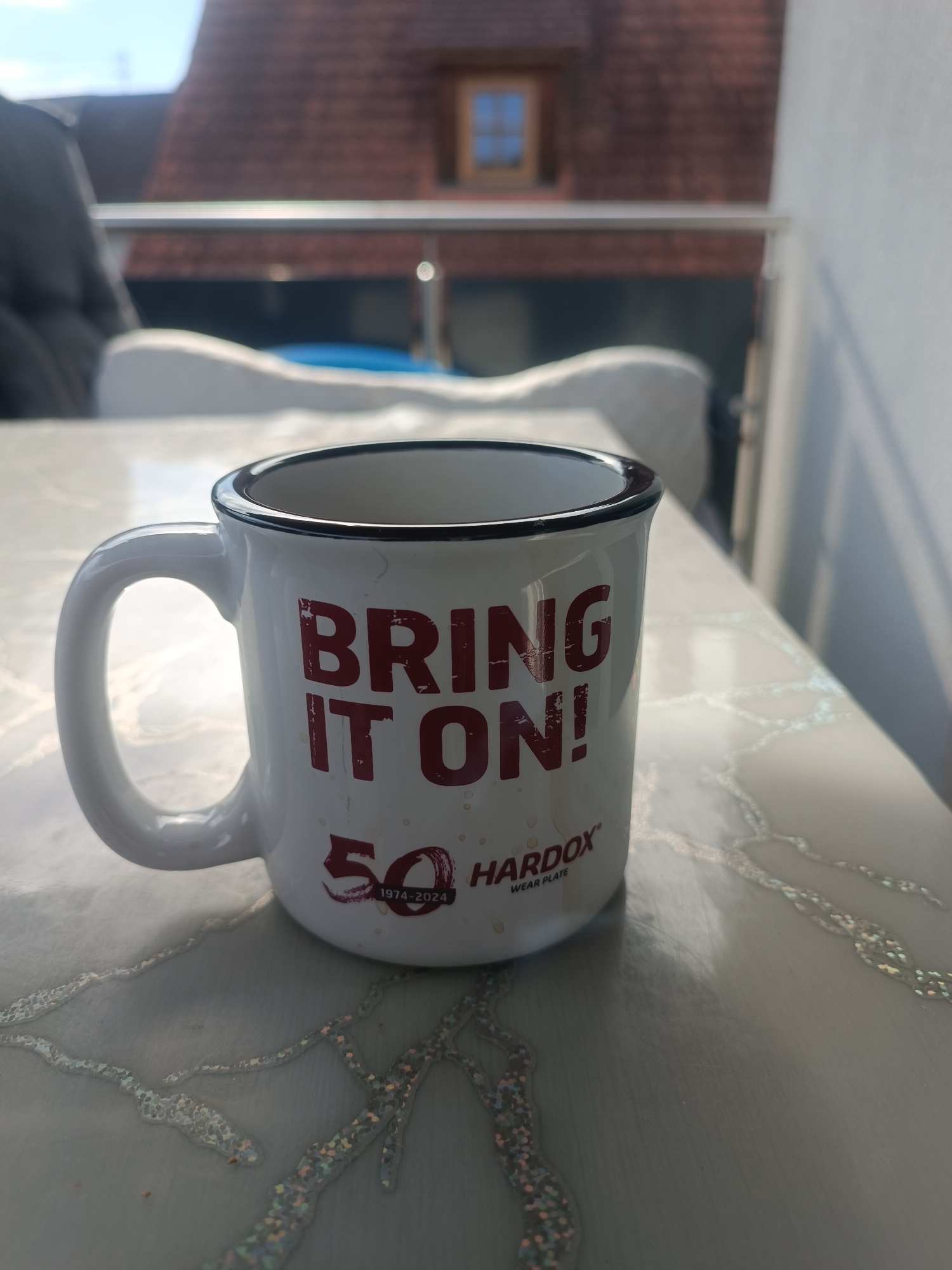 A white mug with a black rim is positioned on a textured, light-colored table, with a balcony railing and a building in the background. The mug features red lettering that reads "BRING IT ON!" and a logo with the number "50" and "1974-2024 HARDOX WEAR PLATE". The mug is slightly angled, with its handle visible on the left. The background shows a blurred view of a rooftop with a dormer window, indicating an urban or suburban setting. The lighting suggests it is daytime, possibly late afternoon given the shadows. The overall scene appears to be an outdoor setting, likely a balcony or patio. The location is identified as Östringen, Germany, although no specific landmarks confirm this from the image alone. There are no people clearly visible or any actions or interactions depicted.
