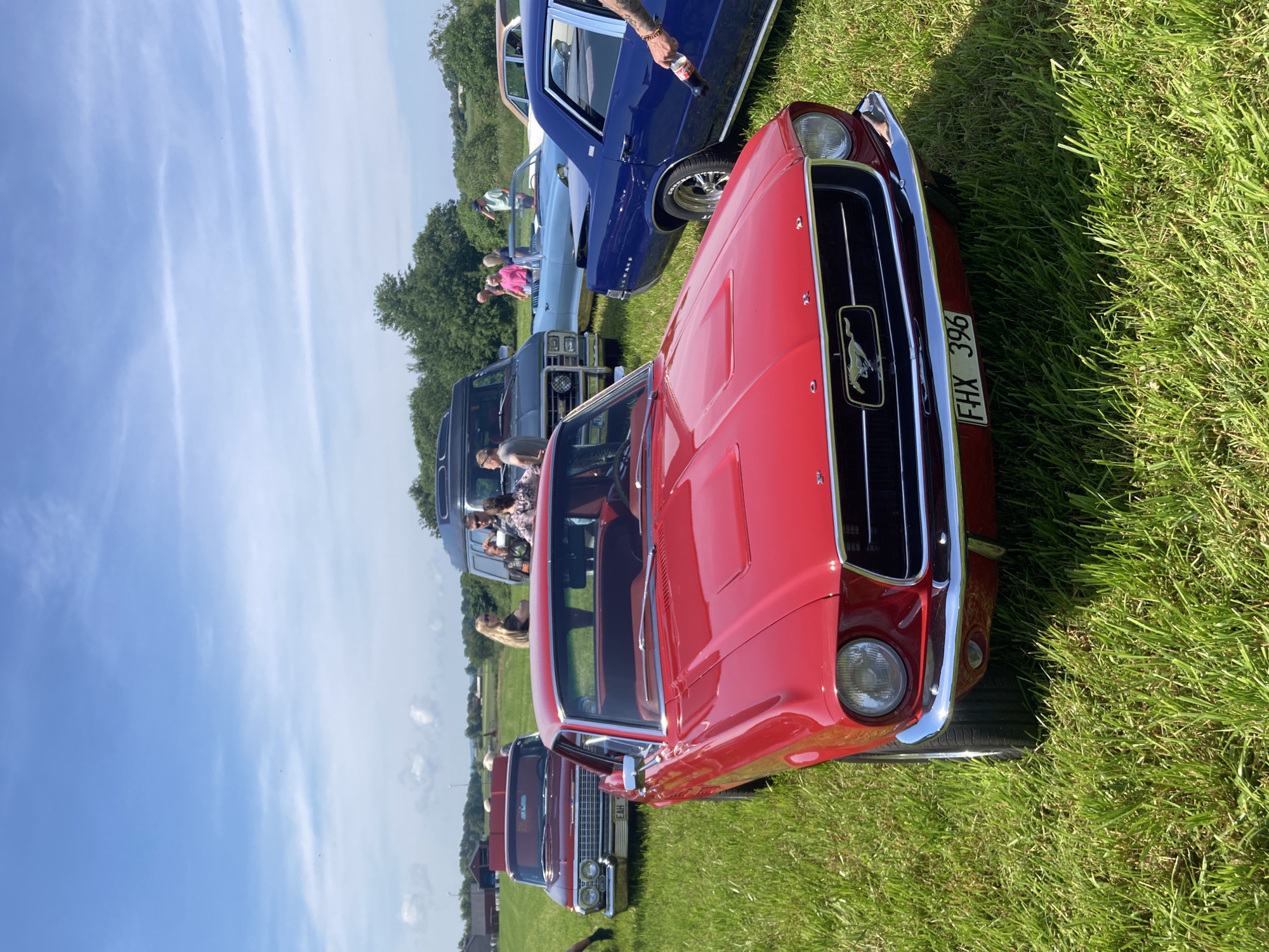 A bright red 1967 Ford Mustang is parked on a grassy field under a partly cloudy blue sky. The Mustang is in the foreground, facing slightly to the left. Its chrome bumper, grille with a pony emblem, and headlights are visible. The license plate reads "FHX 396". Behind the Mustang, other classic cars are parked, including a red Chevrolet Impala to the left, a blue convertible and a dark grey van further back, and a blue sedan on the right. Several people are standing around the vehicles, some conversing and others observing. The environment appears to be an outdoor event, possibly a car show, in a rural setting with trees and fields in the background. The location is identified as Edenberga, Sweden.