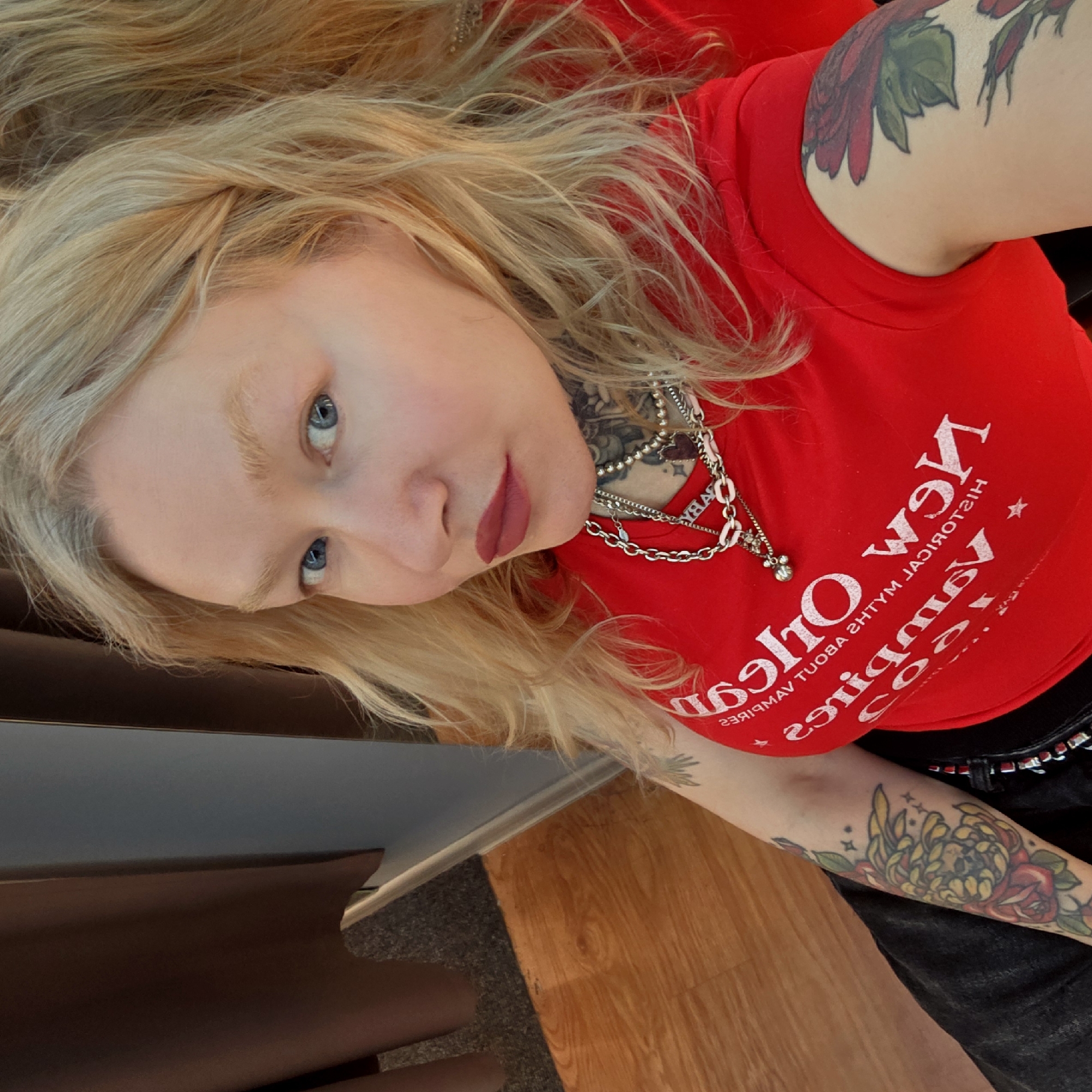 The image features a woman with blonde hair, blue eyes, and light skin, posing indoors in a diagonally tilted frame. She is wearing a red sleeveless top, adorned with multiple silver-toned chain necklaces around her neck and chest. The text on her red top is visible in reverse, reading "New Orleans Historical Myths About Vampires". Her lips are colored with red lipstick.
Visible tattoos include a red and green floral design on her right upper arm (viewer's left), and a colorful floral pattern with prominent yellow, red, and green elements, possibly depicting a chrysanthemum and a rose with small stars, on her left forearm (viewer's right).
The background shows wooden flooring and a carpeted area, suggesting an indoor domestic setting, potentially near stairs. The lighting appears bright, indicating daytime. The woman looks towards the camera with a neutral expression. The location context for this media file is Saint Petersburg, Russia.