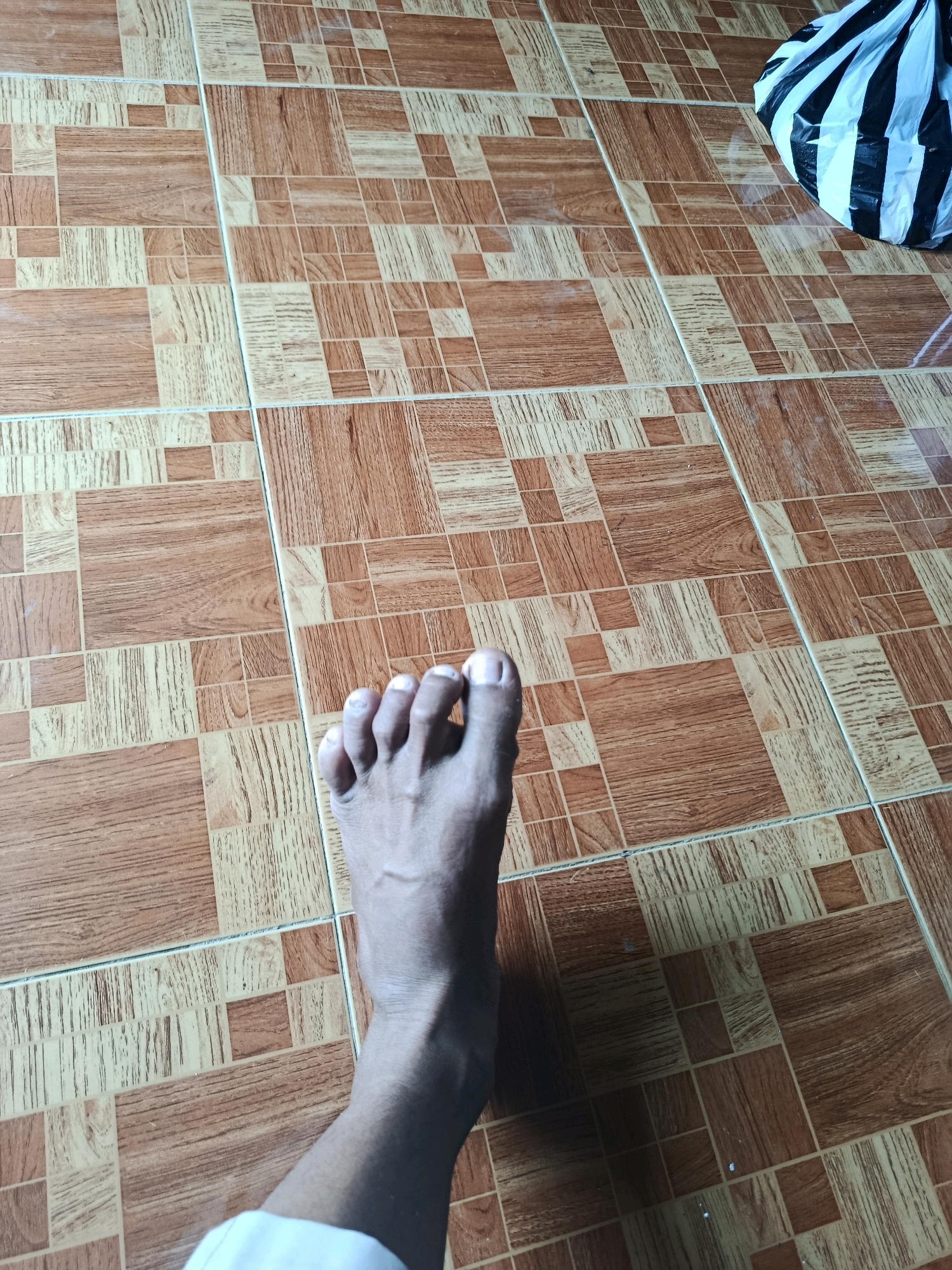An overhead shot captures a person's left foot and ankle positioned on a tiled floor. The tiles are square and arranged in a repeating pattern of light beige and reddish-brown squares, simulating a wood-grain texture. Some tiles are solid colors, while others display distinct grain lines. The foot is bare, with toes slightly splayed, and the skin appears tanned. Veins are visible on the ankle. A white garment is visible at the bottom edge of the frame, partially obscuring the lower leg. In the upper right corner, a portion of a black and white striped plastic bag is visible. The lighting suggests an indoor environment, with a shadow cast by the foot. The location is identified as Binangun, Indonesia.