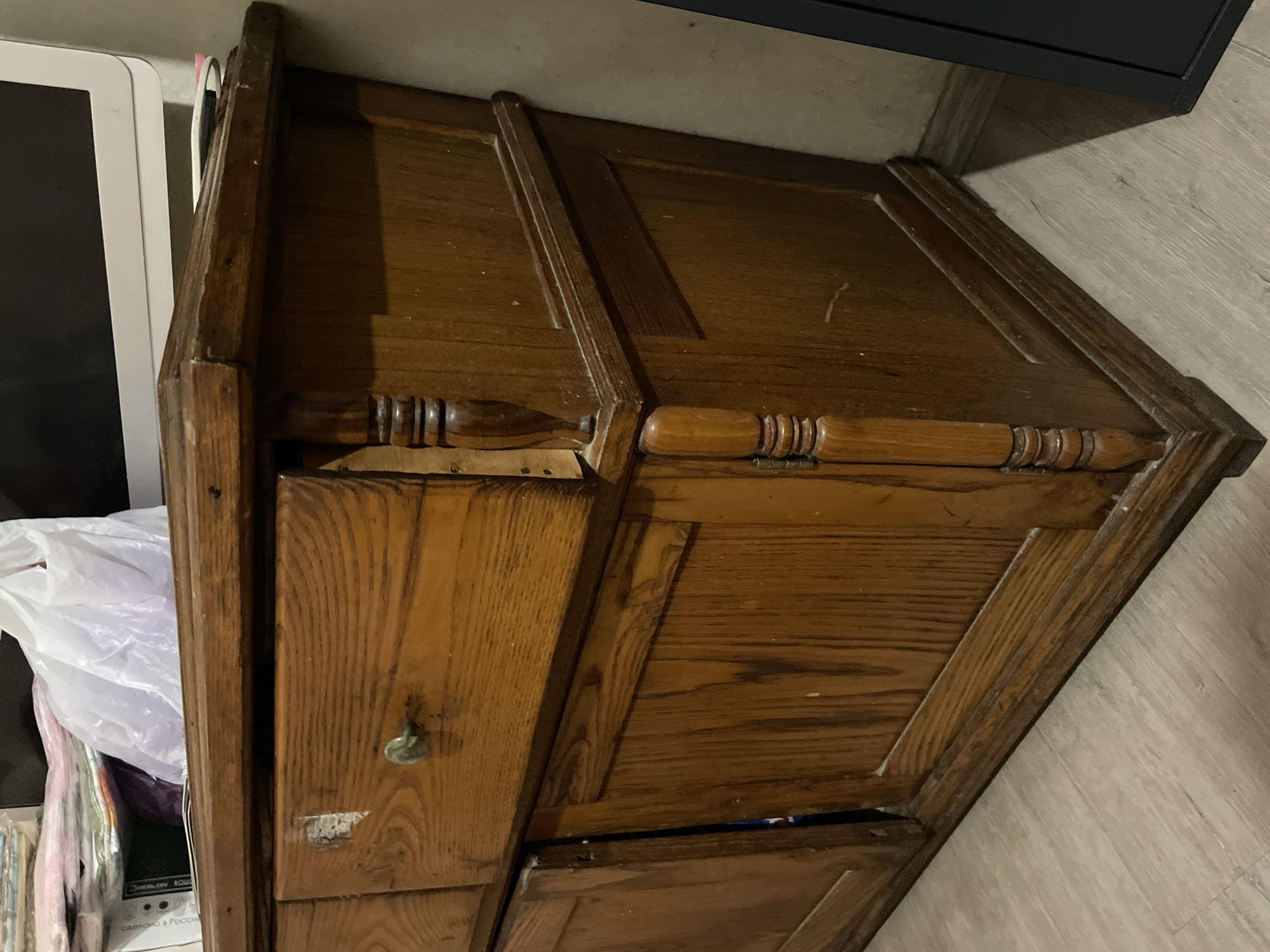 The image shows a wooden cabinet with a drawer, located indoors. The cabinet appears to be old and has a visible keyhole on the drawer. To the left of the cabinet, there is a white object (likely a monitor or screen) and a plastic bag filled with items. The surrounding environment suggests an interior space, possibly a room with light-colored flooring. There is no evidence in the image to support it being located in the city of Olginka, Russia.