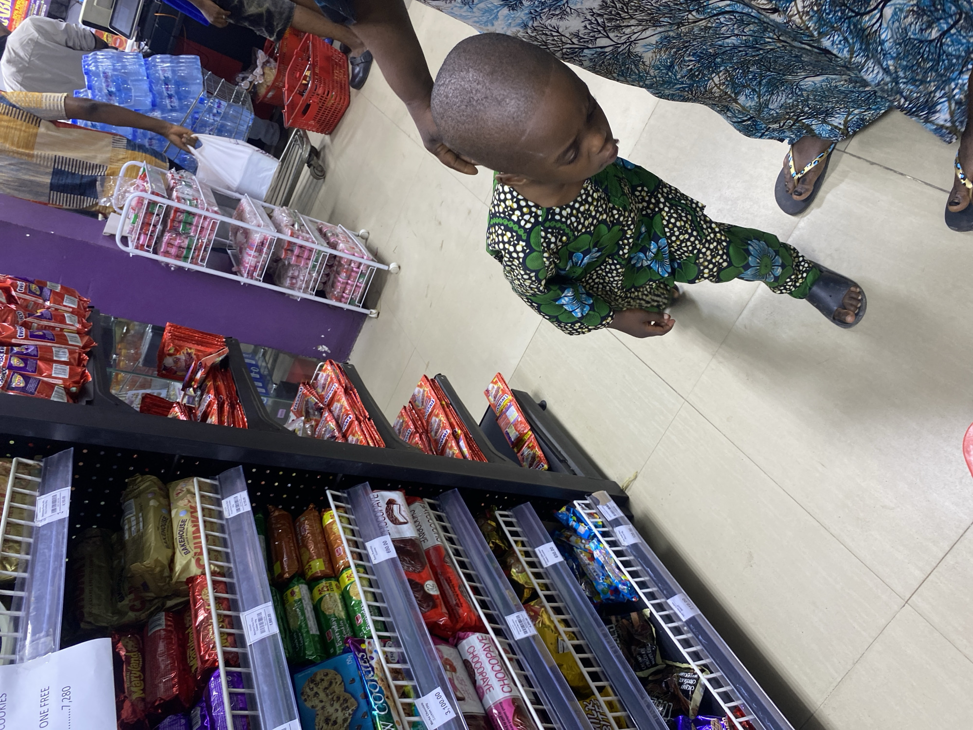 The image shows a scene inside a store, likely a supermarket, in Lagos, Nigeria. A young child, wearing a green and white patterned outfit, is standing and looking at a shelf filled with various snacks, including cookies and chocolate bars. The shelves have clear plastic dividers. Price tags are visible on the shelves. An adult is standing behind the child, presumably accompanying them. The adult is wearing a blue patterned dress and flip-flops. Other individuals are partially visible in the background, including one who appears to be holding bottled water and a red shopping basket. The floor is tiled in a light color. The overall setting suggests a typical shopping environment.