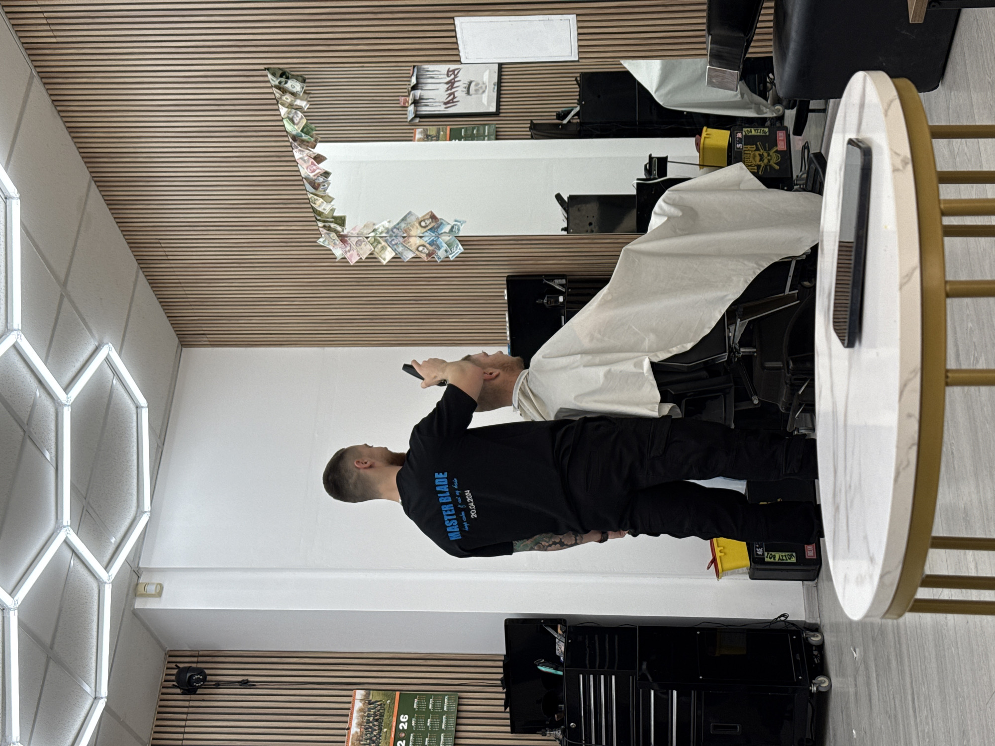 A barbershop scene is depicted. A barber with short hair and a black long-sleeved shirt is standing next to a barber chair, attending to a client with a white smock. The shirt has "MASTER BLADE" printed on the back. The barbershop has a modern interior with a mirror surrounded by a money design, wooden paneling on the walls, and hexagon-shaped lighting fixtures on the ceiling. The scene includes salon equipment like barber chairs and tool cabinets. The location is Sofia, Bulgaria, as indicated by the Bulgarian currency used in the money design around the mirror.