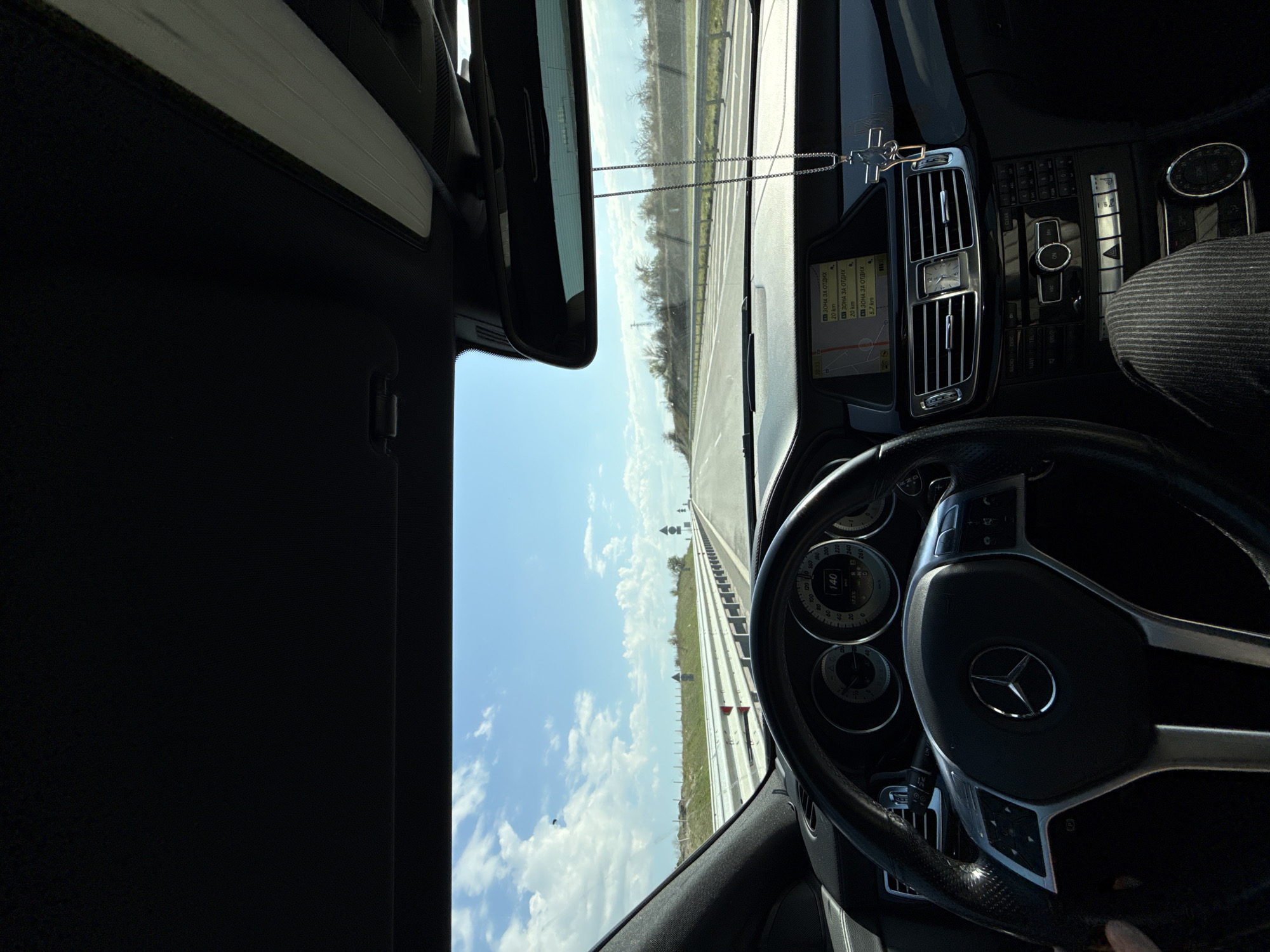 The image depicts the interior of a Mercedes-Benz car, likely while in motion on a highway near the city of Svoboda, Bulgaria. The view is from the driver's perspective, with the steering wheel prominently featuring the Mercedes-Benz logo. The dashboard is visible, showing a navigation screen with text in Bulgarian and some buttons. The speedometer indicates a speed exceeding 88 kilometers per hour. Beyond the windshield, a straight highway stretches into the distance under a blue sky with scattered clouds. The landscape beside the road appears to be a combination of grassy fields and trees. The image orientation is rotated, likely from being taken with a mobile device.