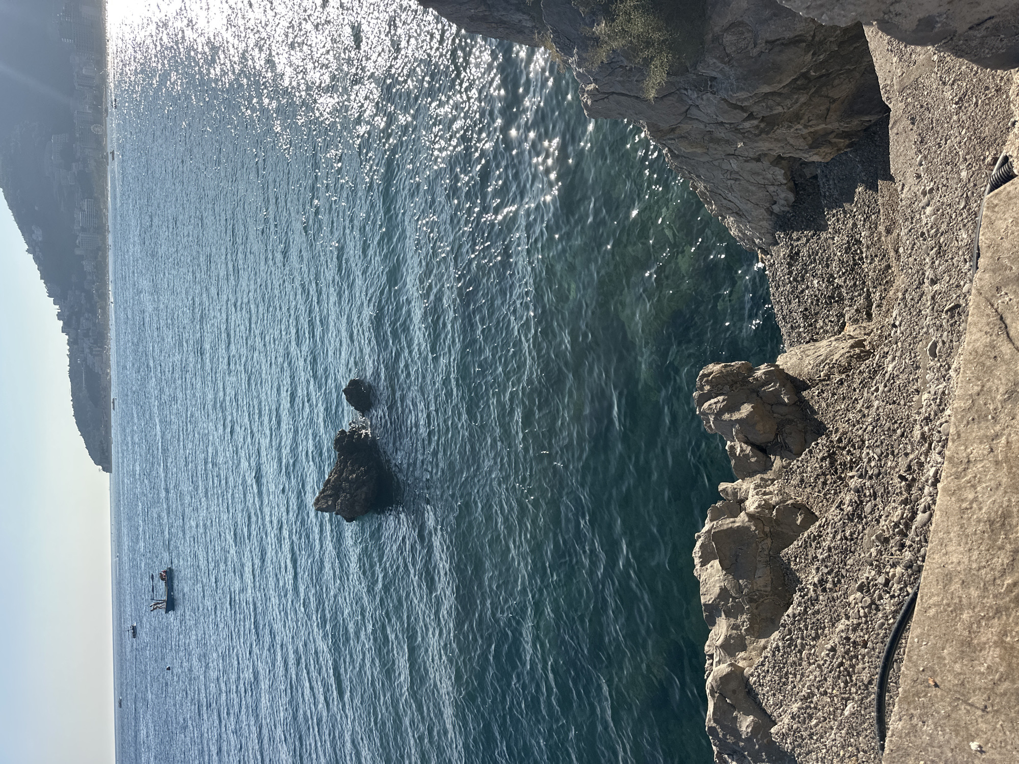 The image shows a view of the Adriatic Sea with the coastline of Budva, Montenegro, in the background. The foreground features a rocky shoreline with a concrete pathway and steps leading down towards the water. The sea is a deep blue-green, with sunlight glinting off the surface. In the middle distance, two dark rocks emerge from the water. Further out, a small boat with several people on board is visible. The distant coastline is lined with buildings and a green, mountainous landscape rises behind them. The sky is a clear, pale blue. The scene appears to be a scenic overlook or a small harbor area.