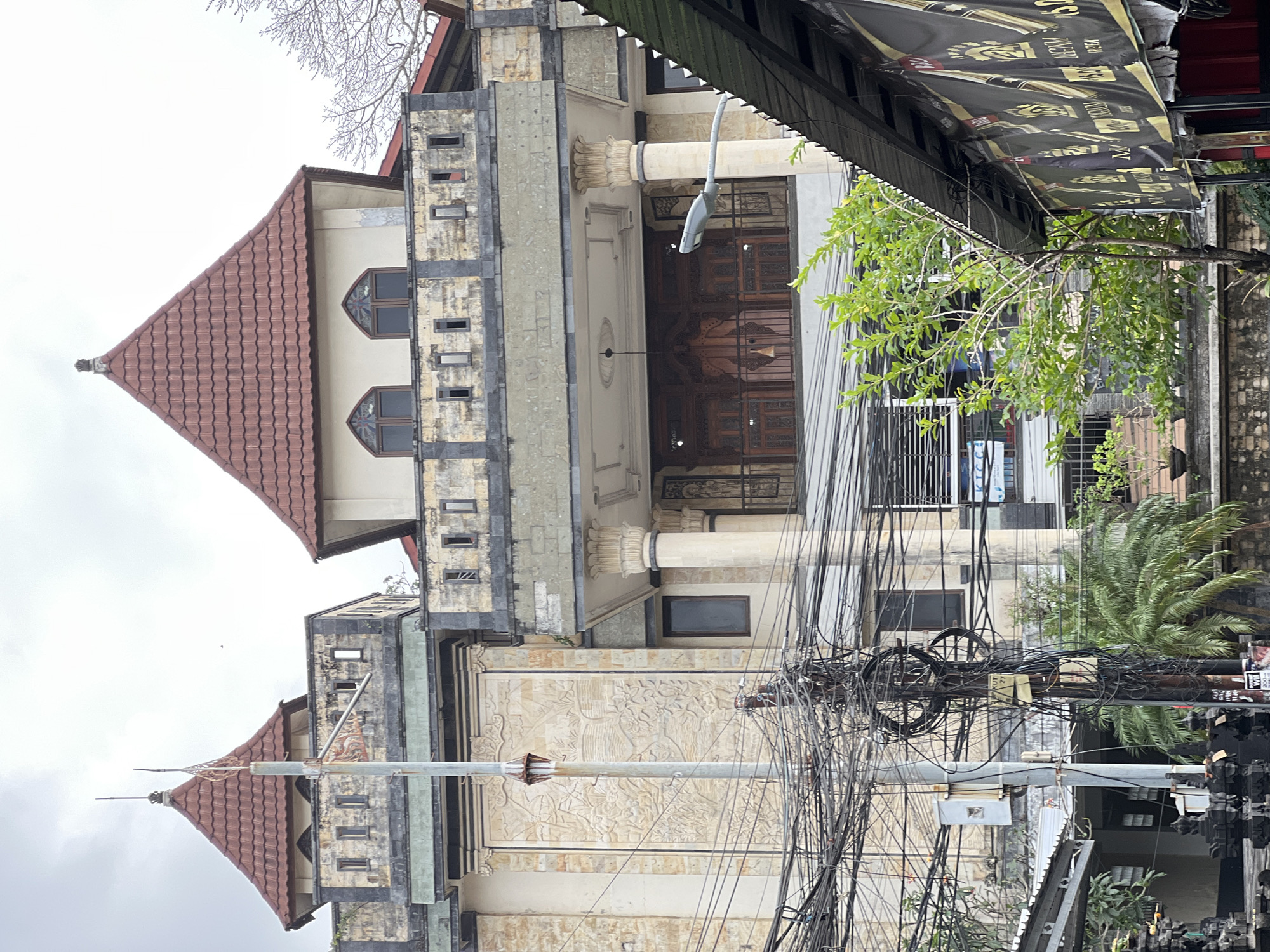 A street-level, eye-level, medium shot captures a multi-story building with two distinct turret-like structures, each topped with a red-tiled pyramid roof. The building's facade is a mix of textured beige stone and lighter-colored plaster. The lower levels feature ornate carvings on a large beige panel, flanked by smaller rectangular windows. A covered balcony with decorative columns and intricate wooden doors is visible on the second floor. The foreground is dominated by a chaotic tangle of electrical wires and a utility pole, obstructing the view of the building's lower section. A street light extends from the right side of the image, pointing towards the building. Lush green trees and foliage grow in front of and around the building. A dark-colored awning with gold lettering is visible on the far right, suggesting a commercial establishment. The sky is overcast and light gray. The scene appears to be a street in Kuta, Indonesia, characterized by dense electrical infrastructure and tropical vegetation. No people are visible in the image.