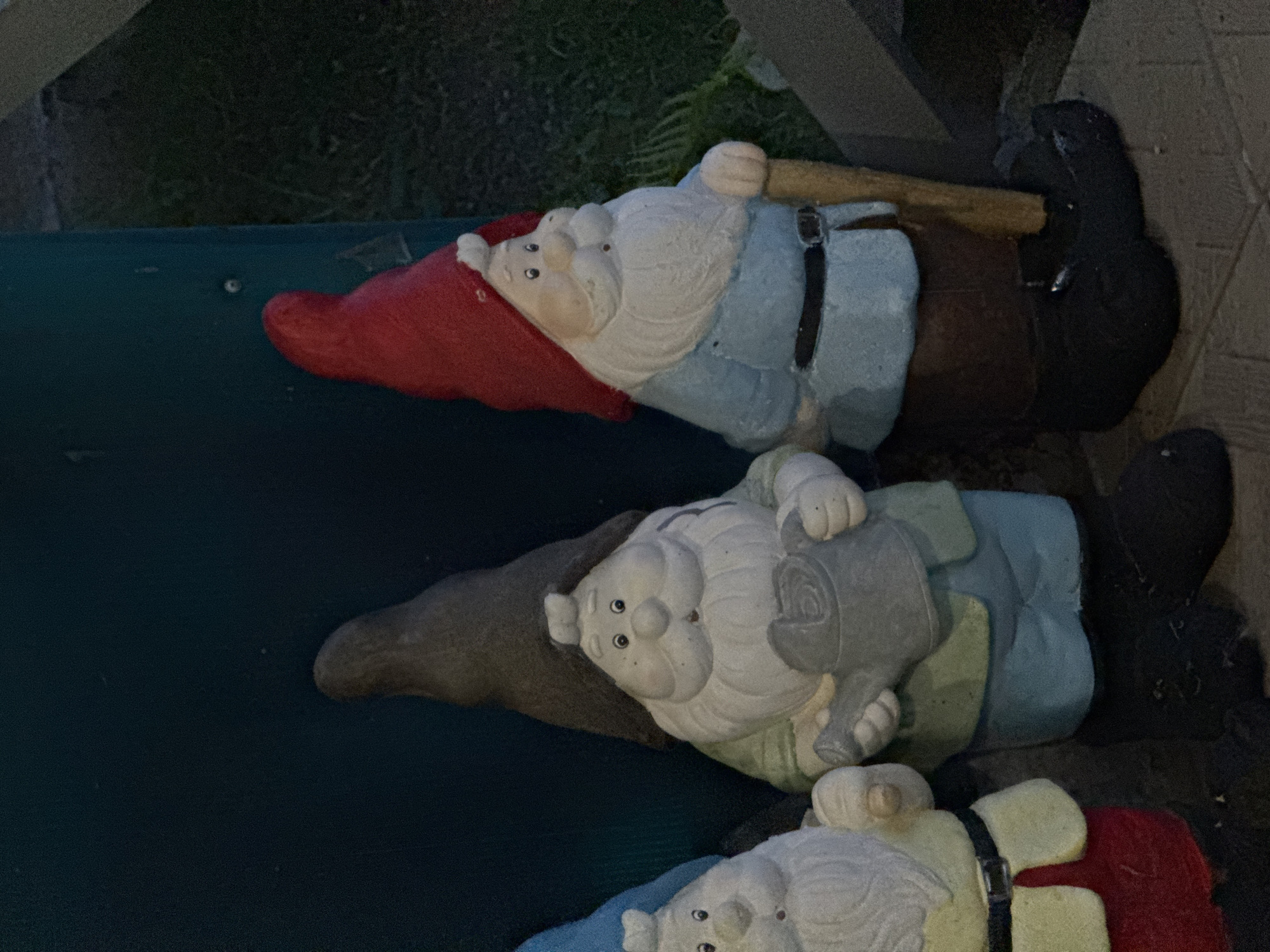 Three garden gnome statues are arranged in a row. The leftmost gnome has a blue hat and a cream-colored beard, wearing a yellow jacket over a red lower garment and a black belt. The middle gnome wears a dark gray pointed hat, a cream-colored beard, a light green tunic, light blue pants, and is holding a watering can. The rightmost gnome has a red pointed hat, a cream-colored beard, a light blue tunic, brown pants, a black belt, and is holding a wooden staff. The statues are positioned outdoors, on a paved surface, with a dark green background and some greenery visible to the right. The image appears to have been taken in Sakmara, Russia, likely in a garden or yard setting.