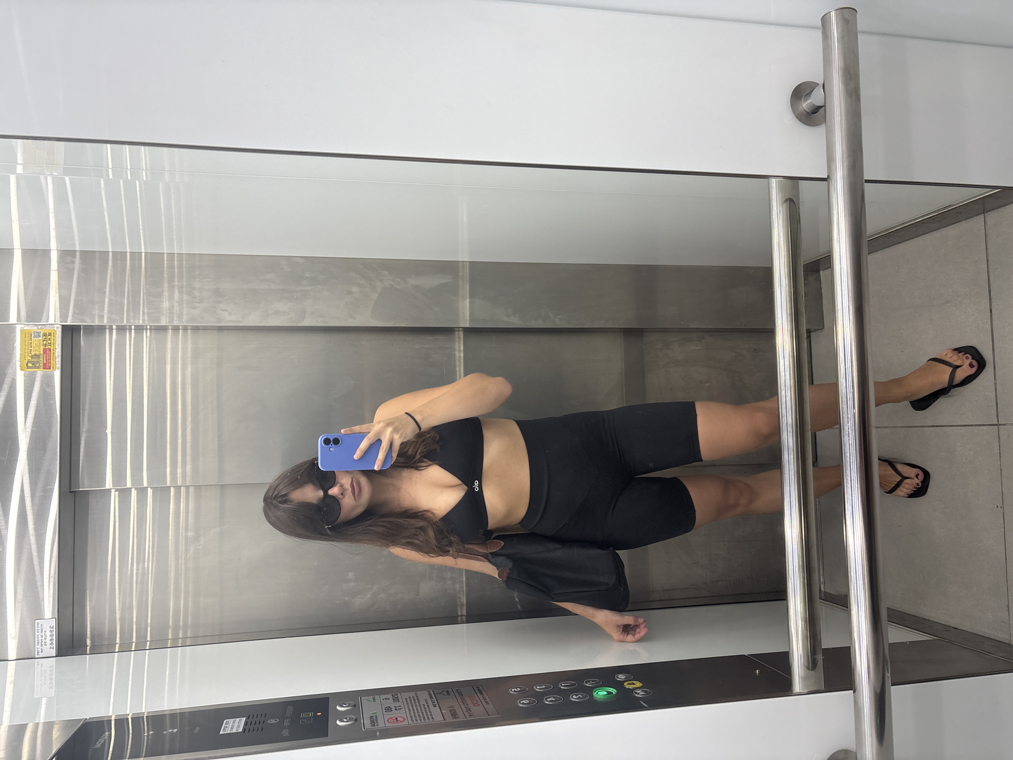 A woman with brown hair and sunglasses is standing in an elevator, taking a mirror selfie. She is wearing a black sports bra and black biker shorts, and she has a black tote bag slung over her left shoulder. She is also wearing black flip-flops. The elevator walls are made of brushed metal, and the floor is tiled. There is an elevator control panel on the left side of the elevator, with several buttons, one of which is illuminated green. A sign is visible above the control panel. The elevator appears to be located in Tel-Aviv, Israel, based on information associated with the image.