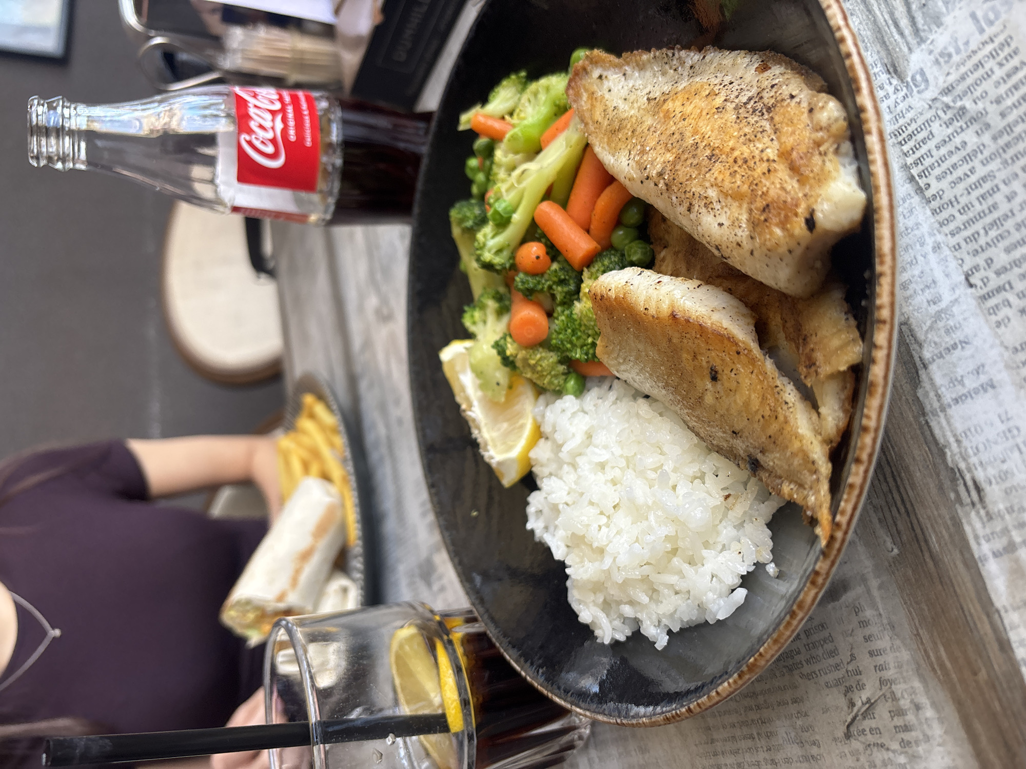 The image shows a meal served at a table in Plovdiv, Bulgaria. The main dish consists of pan-fried fish fillets, white rice, steamed broccoli, carrots, and peas, garnished with a lemon wedge, presented on a dark-colored plate. A bottle of Coca-Cola, a glass with a straw and a lemon slice, and what appears to be a wrap and fries are also visible on the table. A person with a dark top is partially visible in the lower left corner of the frame. The table has a newspaper print design on its surface.