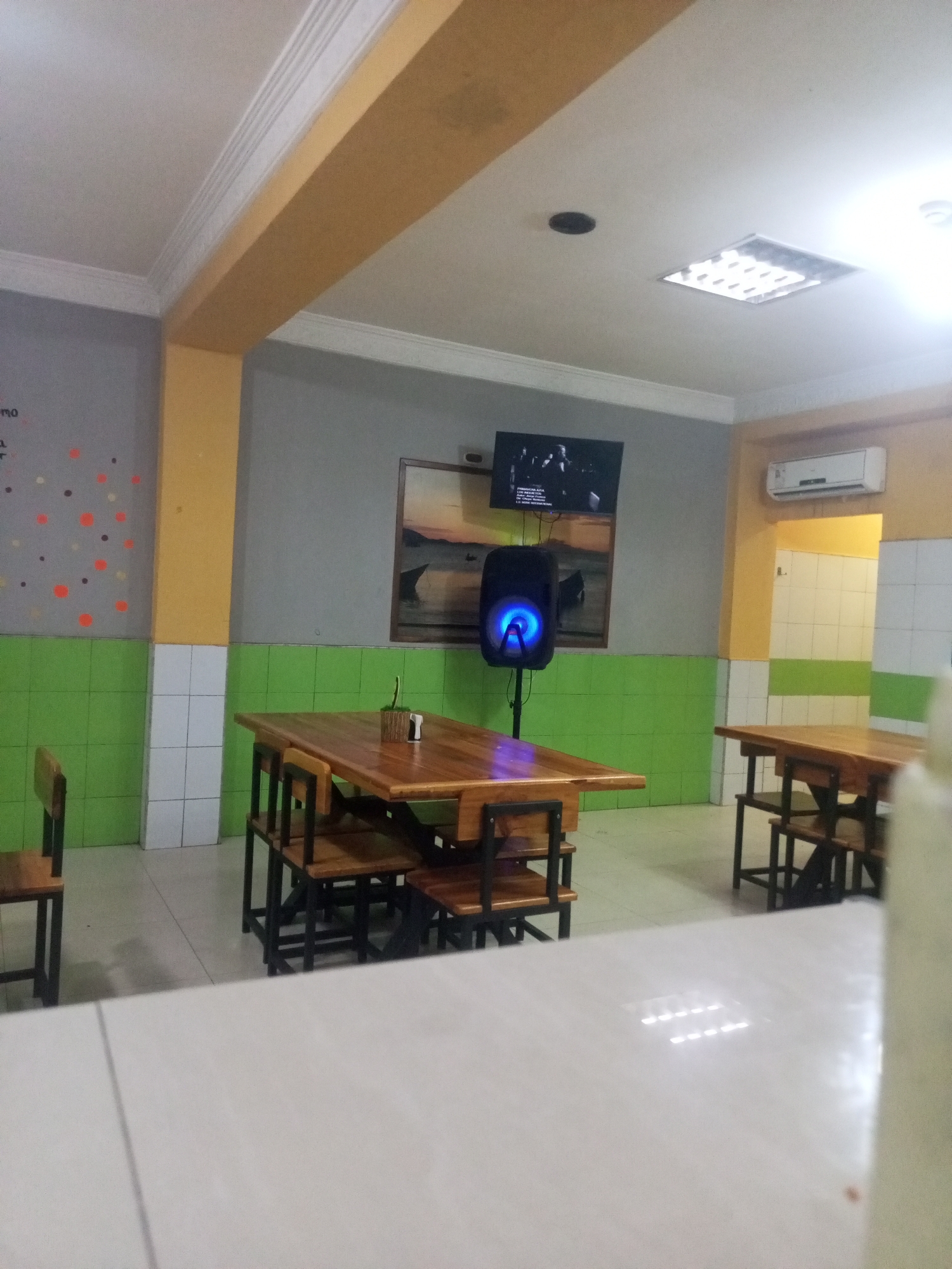The image depicts an interior space in what appears to be a restaurant or cafeteria in Juan Griego, Venezuela. The room is characterized by a color scheme of grey, yellow, white, and green, with tiled walls and a drop ceiling. Three wooden tables with attached wooden chairs are visible, two of which are in focus while the third is partially obscured by the foreground. A television is mounted on the wall, displaying a monochrome video. Below the television, a blue-lit speaker stands on a tripod. A painting hangs on the wall beside the speaker. A section of the wall is decorated with painted circles. An air conditioning unit is visible above a doorway. No people are clearly discernible in the image. The foreground is occupied by a white tabletop.