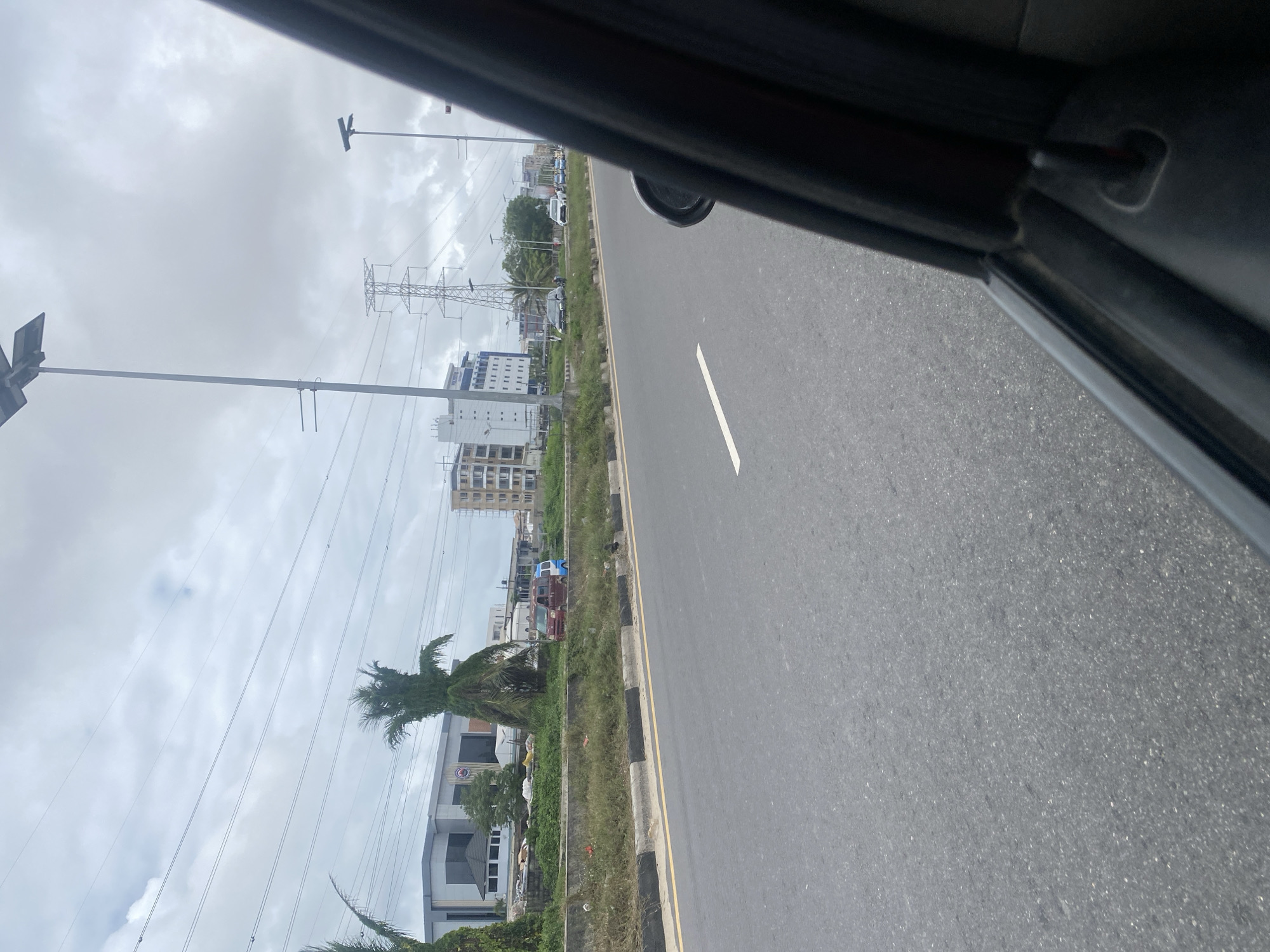 The image captures a view from a vehicle in Ilado, Nigeria, facing forward on a paved road. The road has a yellow and black striped median and a white line marking. On the left side of the road, there is a grassy median with some debris and vegetation. Behind the median, a modern building with a glass facade and a peaked roof is visible, along with some palm trees. Further in the distance, there are tall power lines, electrical towers, and several multi-story buildings. A few vehicles, including a red van and a blue and white van, are parked or driving on the road. The sky is overcast with clouds. The right side of the image shows the interior of the vehicle, specifically the door frame and a portion of the passenger seat.