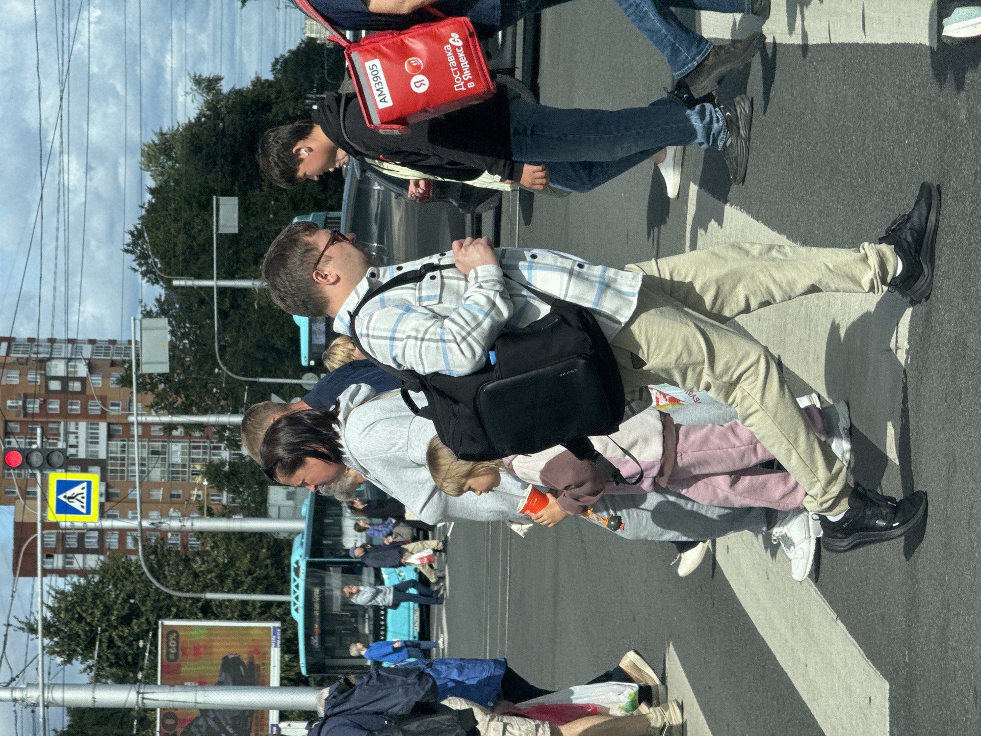In Ladozhskoe Ozero, Russia, a pedestrian crossing is depicted on a sunny day. A group of people are crossing the street. A man in a plaid shirt and beige pants, carrying a black backpack, is walking across the crosswalk. To his right, a young man wearing a black hoodie and blue jeans is also crossing, with a red delivery bag labeled "AM3905 Доставка в Яндекс.Go" on his back. A woman in a grey hoodie and a young girl in a pink and beige outfit are walking together near the man with the plaid shirt. In the background, a light blue bus with "VOLGABUS" written on it is stopped, and several other pedestrians are visible. Traffic lights and street signs, including a pedestrian crossing sign, are present. Buildings and trees form the backdrop.