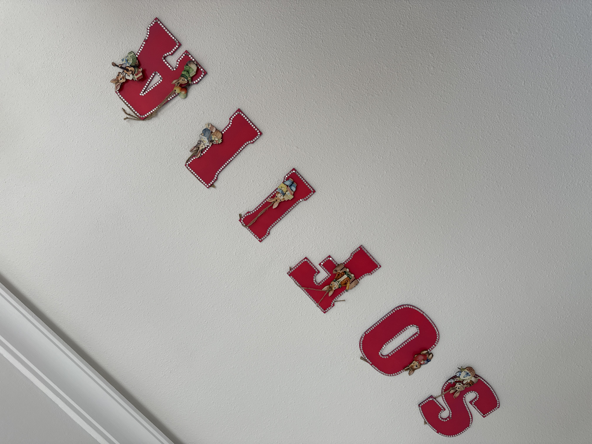 The image shows a wall in a room, likely a child's room, with the name "KELSEY" spelled out vertically using red, decorated letters. Each letter appears to have a border of small, shiny embellishments, possibly rhinestones. In addition, each letter is adorned with vintage-style illustrations of animals, such as rabbits. The wall is a light gray or white color with a lightly textured surface. A section of white molding, featuring two recessed parallel lines, is visible in the lower left corner of the image. The letters are positioned as if hanging on the wall. The room appears to be in Unknown, Unknown.