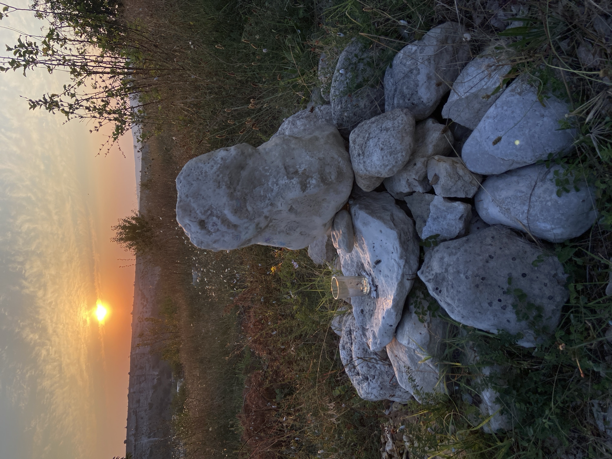The image captures a sunset over a natural landscape, likely in Krasnomakskoye Rural Settlement, Ukraine. In the foreground, a pile of white and grey stones is arranged, with a small glass and some pebbles placed on one of the stones. The stones vary in size and texture. Behind the stones, dry grasses and some green foliage create a rugged terrain. In the middle ground, a gentle slope leads down to a valley with a distant cliff face. The sky is dominated by the warm hues of the setting sun, with scattered clouds tinged with orange and yellow. Silhouettes of trees and shrubs are visible against the horizon. The overall lighting suggests the golden hour, casting long shadows and illuminating the scene with a soft glow.