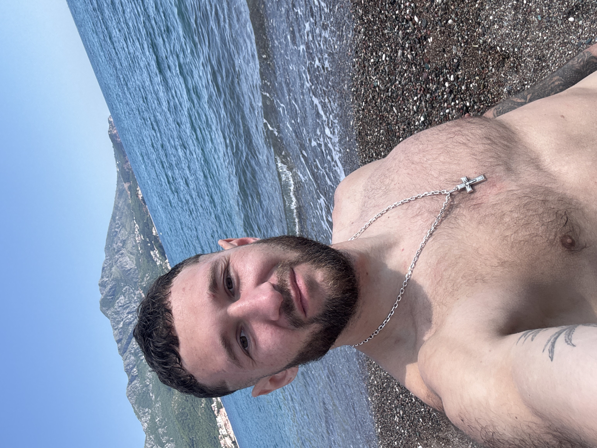 A man with a beard, wearing a silver cross necklace, is at a beach in the city of Unknown, Unknown. The beach has dark pebbles. A mountain is visible in the background. The water is light blue with white waves. The sky is a clear, light blue.