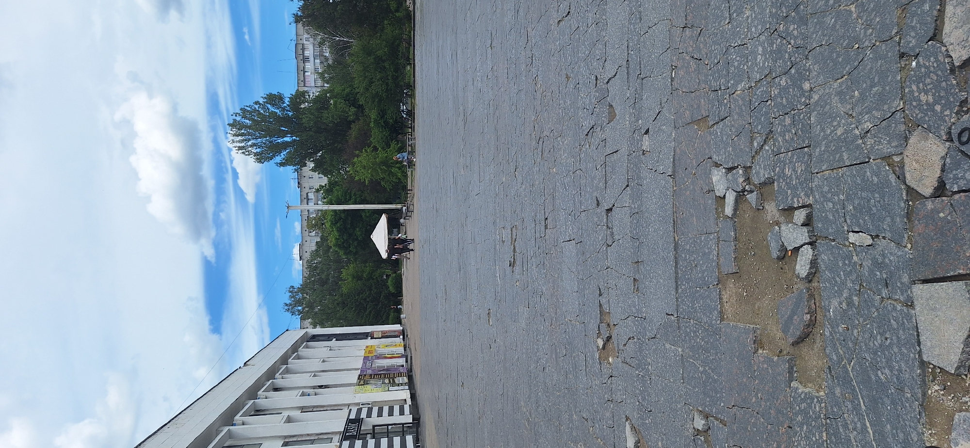 The image depicts an outdoor scene in the city of Bila Tserkva, Ukraine, likely a public square or plaza. In the foreground, the ground is paved with large, dark grey stone slabs that are cracked and broken in several places, with some areas showing exposed dirt and debris.
To the left, a long, white building with a modern facade is visible. It features a row of vertical columns and appears to house multiple businesses or shops, with signage including "BASIC" on one of them.
In the middle ground, a white canopy tent is set up, and several people are walking or standing near it. Beyond the tent, a variety of trees and greenery form a dense line, with taller trees in the background. Further back, multi-story apartment buildings are visible, characteristic of urban landscapes.
A single streetlamp stands tall amidst the trees, and a few park benches are scattered in the area. A person is seated on one of the benches in the right middle ground. The sky above is partly cloudy, with patches of blue sky interspersed with white clouds. The overall impression is one of a somewhat worn but functional public space on a bright day.