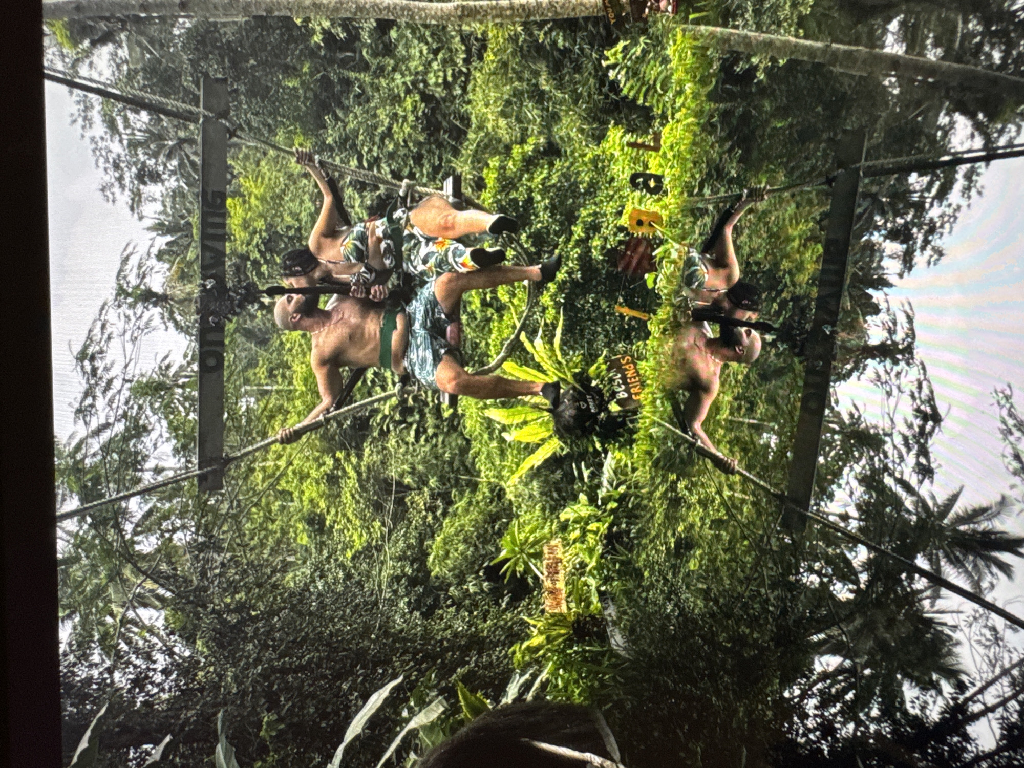 Two shirtless men are performing on a high ropes course in a lush jungle environment. One man is holding onto the rope with his arms outstretched, while the other man is sitting on his shoulders. Both men are wearing shorts. The background is filled with dense green vegetation. The location is in the city of Unknown, Unknown.