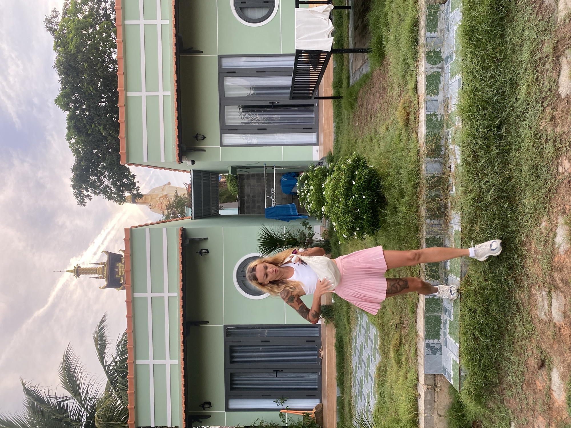 In Mũi Né, Vietnam, a woman walks outdoors in front of two identical green buildings with orange tile roofs. She wears a white crop top, a pink pleated skirt, and white sneakers, and carries a white purse. The buildings have large glass doors and small windows. Behind the buildings, a tall statue of a woman holding a child and a golden pagoda are visible against a cloudy sky. A palm tree is on the left side of the frame, and lush green vegetation surrounds the buildings, with a clothesline holding laundry on the right. The ground is covered in grass and dirt.