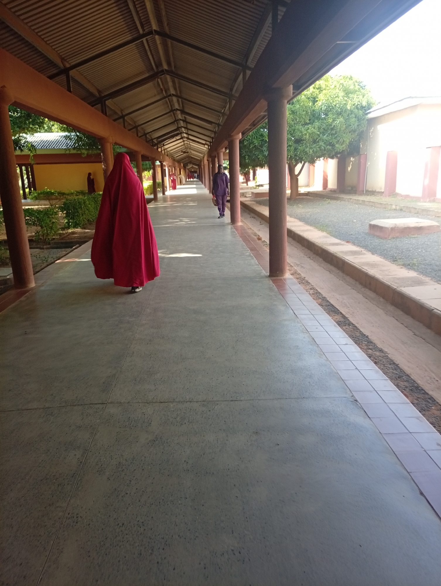 A long, covered walkway with a corrugated metal roof supported by brown pillars stretches into the distance. The walkway is paved with concrete. On the left side, there are yellow buildings with red accents and green shrubs. On the right side, there are beige buildings with red accents and trees. A person in a red, flowing garment is walking away from the camera in the foreground on the left side of the walkway. Further down the walkway, another person in a purple outfit is walking towards the camera. The ground on the right side of the walkway is covered with gravel and there are some concrete structures. The overall impression is of an outdoor school or institutional setting. This scene is likely located in the city of Gombe, Nigeria, given the architectural style and vegetation.