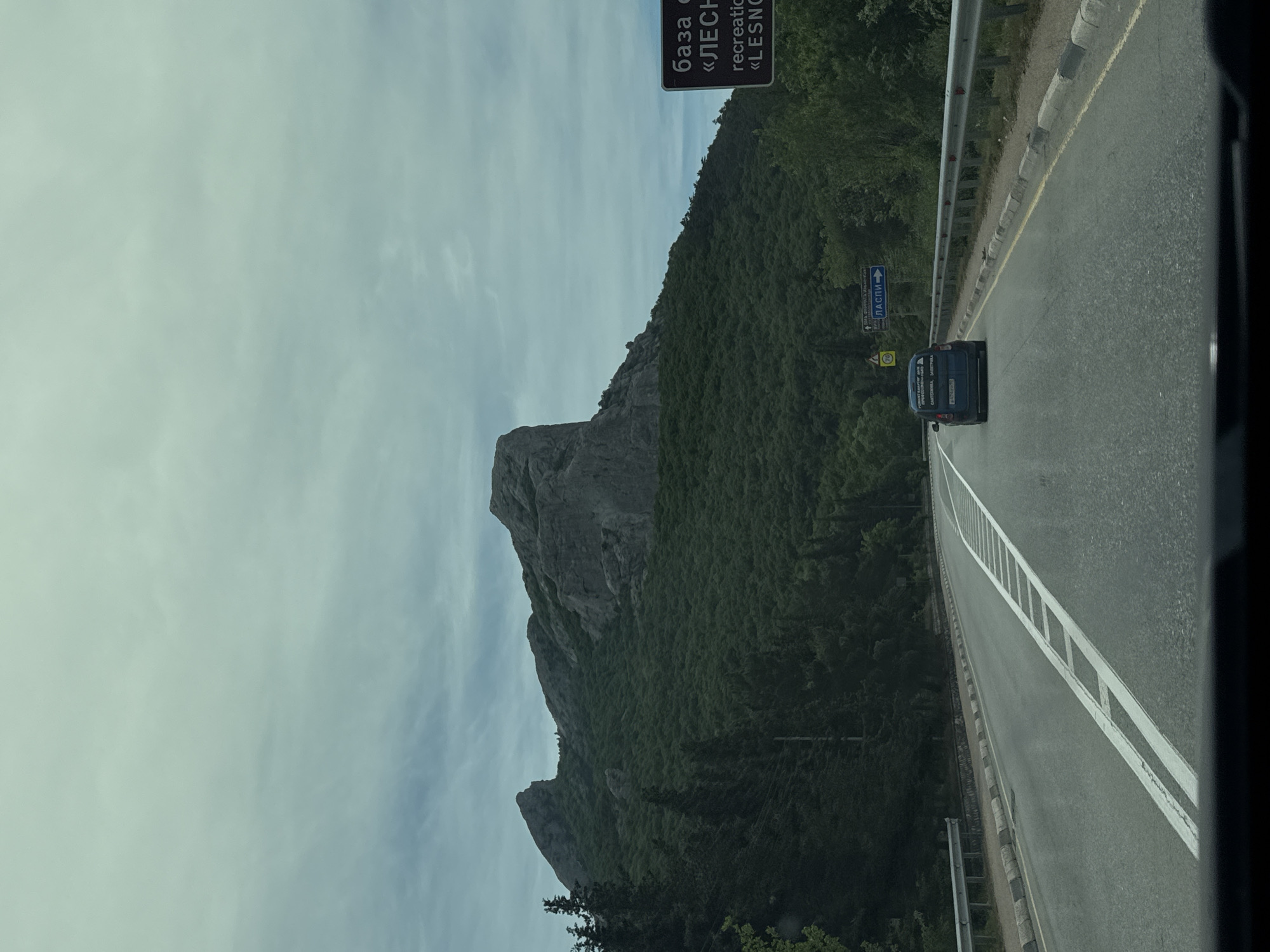 The image shows a road in the Орлиновский округ, Ukraine, with a large rock formation on the left side. A dark-colored car is traveling on the road. There are signs visible along the side of the road, with lettering in Cyrillic. The environment is forested with a cloudy sky above.