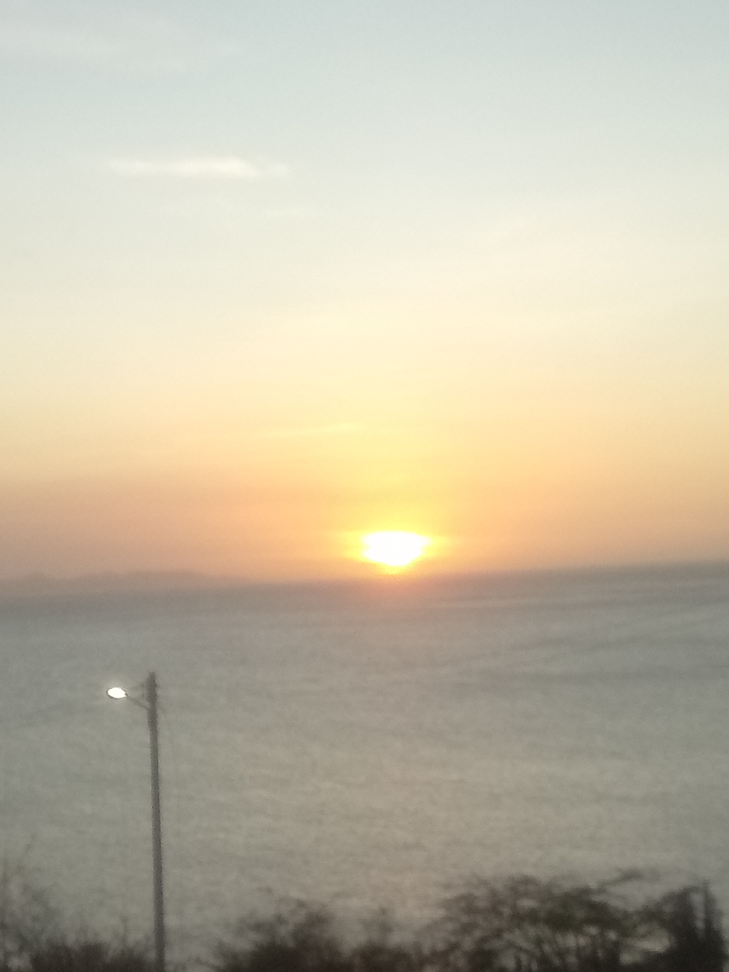 The scene captures a coastal view at either sunrise or sunset over the Caribbean Sea, located in Juan Griego, Venezuela. The sky, a gradient from pale blue at the top to bright orange-yellow near the horizon, occupies the upper two-thirds of the image. The sun, a partially obscured, intensely bright yellow orb, is situated directly on the horizon, slightly right of the vertical center. A faint, wispy cloud is discernible in the upper-mid portion of the sky. The water surface, appearing grey-blue and calm, extends across the lower half of the image. A hazy, dark landmass is faintly visible on the distant horizon, positioned to the left of the sun. In the immediate foreground, on the left side, a dark utility pole stands vertically, topped with a single, illuminated white streetlamp. Indistinct dark vegetation is present along the very bottom edge of the image. No individuals or specific activities are observable.