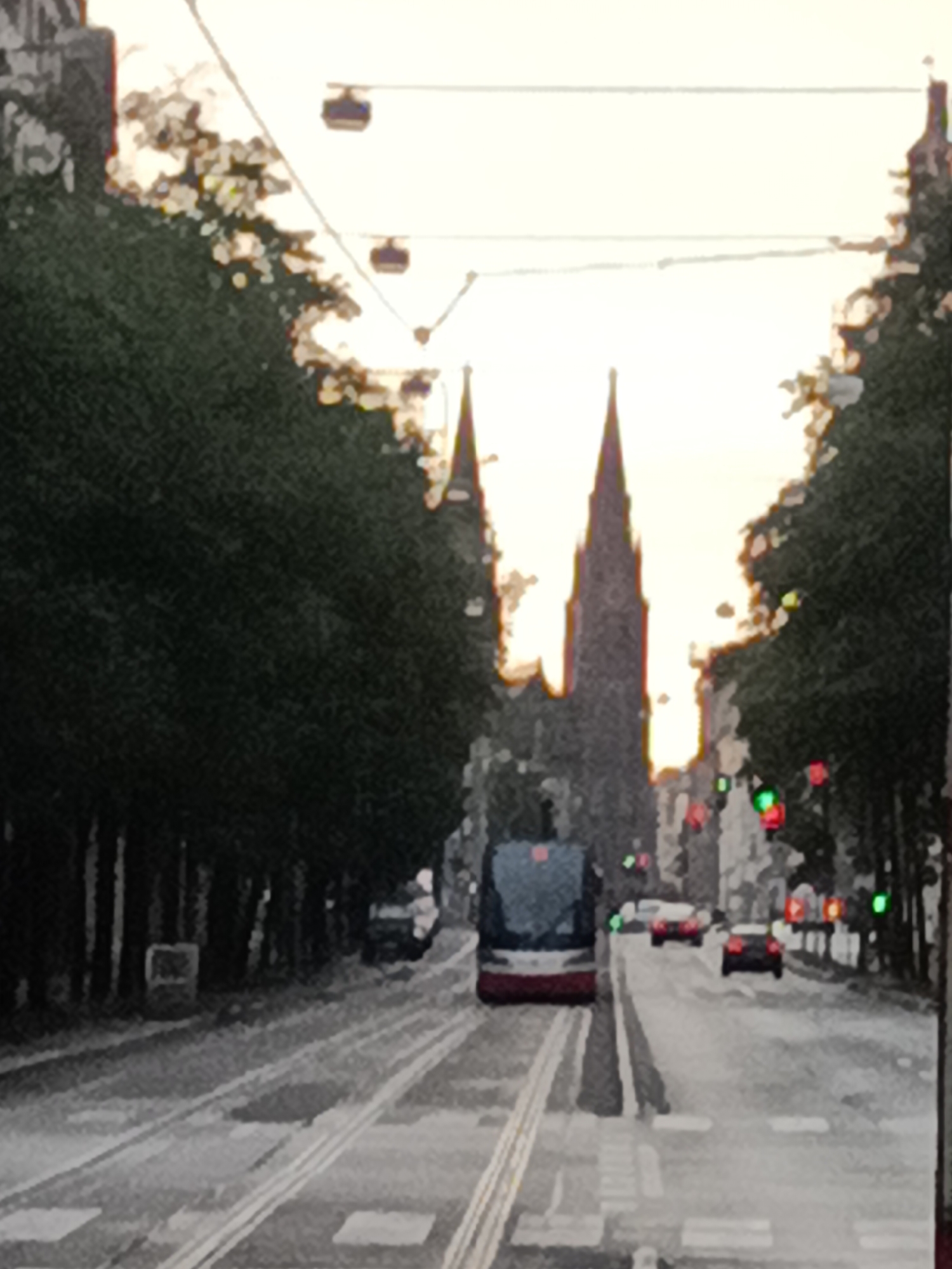 This image captures a street scene in Prague, Czechia. The perspective appears to be from a moving tram, as indicated by the parallel tram tracks in the foreground and the tram itself positioned to the left of the frame. The tram, with its red and white livery, is facing away from the viewer and traveling down the street.
Flanking the tram tracks are rows of mature trees, their dark foliage creating a dramatic contrast with the bright, overcast sky. In the background, two prominent Gothic spires rise above the trees, suggesting the presence of a significant church or cathedral, characteristic of Prague's architecture. Cable car or tramway lines are visible spanning the sky above the street.
Several cars are visible on the road ahead of the tram, including a red car and a dark-colored car, suggesting active traffic. Traffic lights, some displaying green and red signals, are positioned along the street. The overall impression is of a city street during daytime, possibly in the early morning or late afternoon given the diffused light. The image has a slightly soft focus or artistic filter applied, giving it a painterly quality.