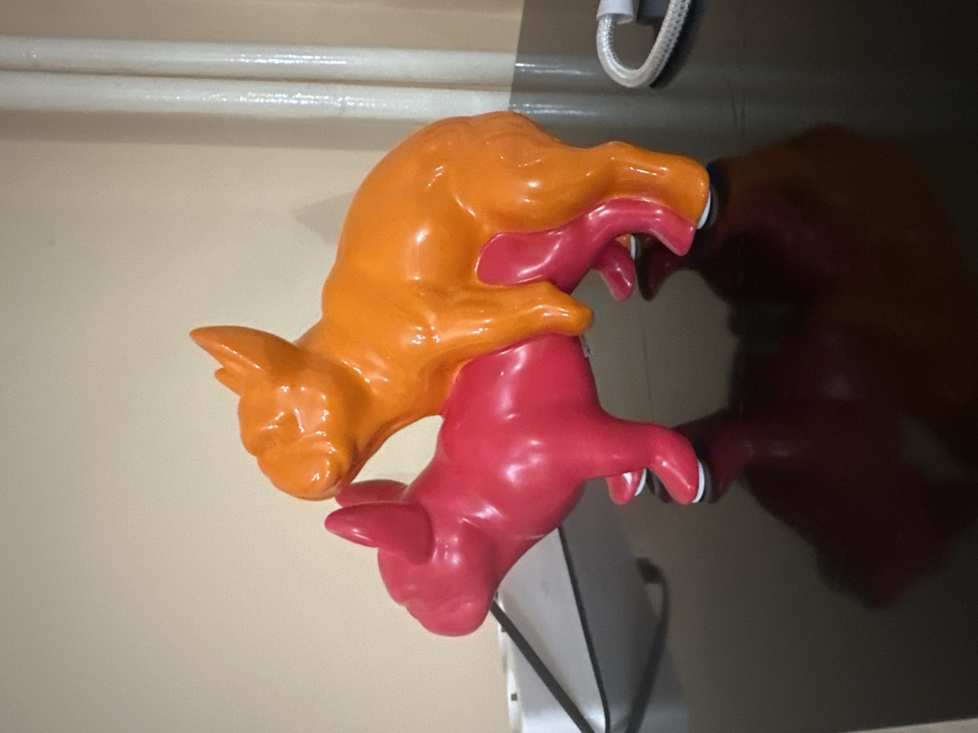 On a surface, likely a television screen given the reflection and presence of a power cord, there are two conjoined pig figurines. One pig is orange and positioned on top of the other, which is pink. The background includes a beige wall and a white horizontal bar. The location is unknown.
