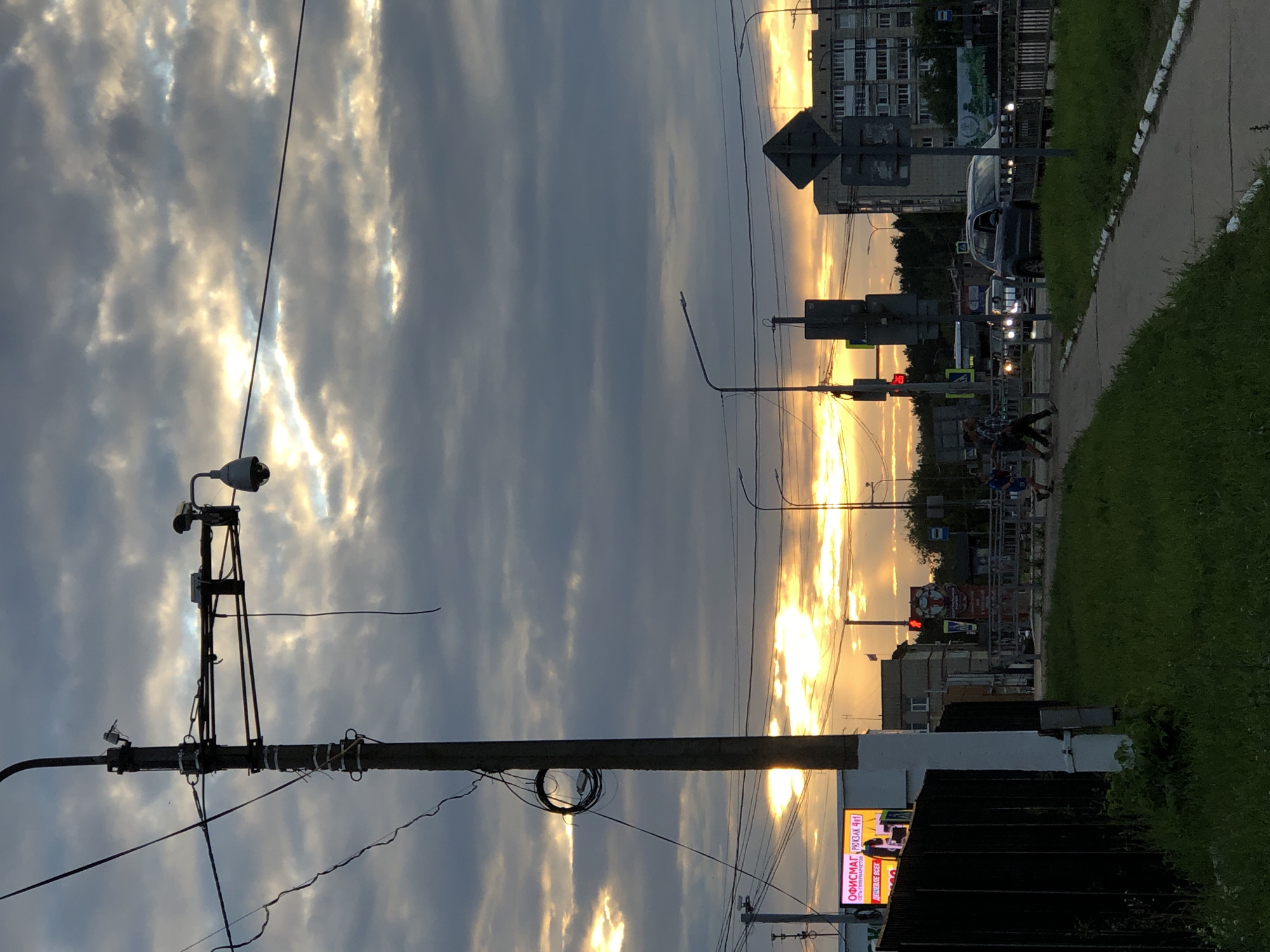 The image captures a street scene in Chaikovsky, Russia, during a cloudy sunset. A tall, weathered utility pole dominates the left foreground, adorned with various wires and two surveillance cameras. To the left of the pole, a black metal fence encloses a grassy area. Beyond the fence, a billboard displays Russian text, including "ОФИСМАГ" and "РЮКЗАК 4 В 1". In the mid-ground, a paved walkway runs alongside a grassy verge. Several cars are visible on a road in the distance, with headlights illuminated. A pedestrian signal displays a red light. Buildings, including a multi-story apartment complex, are situated further back. Three individuals are walking on the sidewalk in the mid-ground. The sky is filled with dramatic clouds, illuminated by the setting sun which creates bright yellow and orange hues in the upper portion of the image.