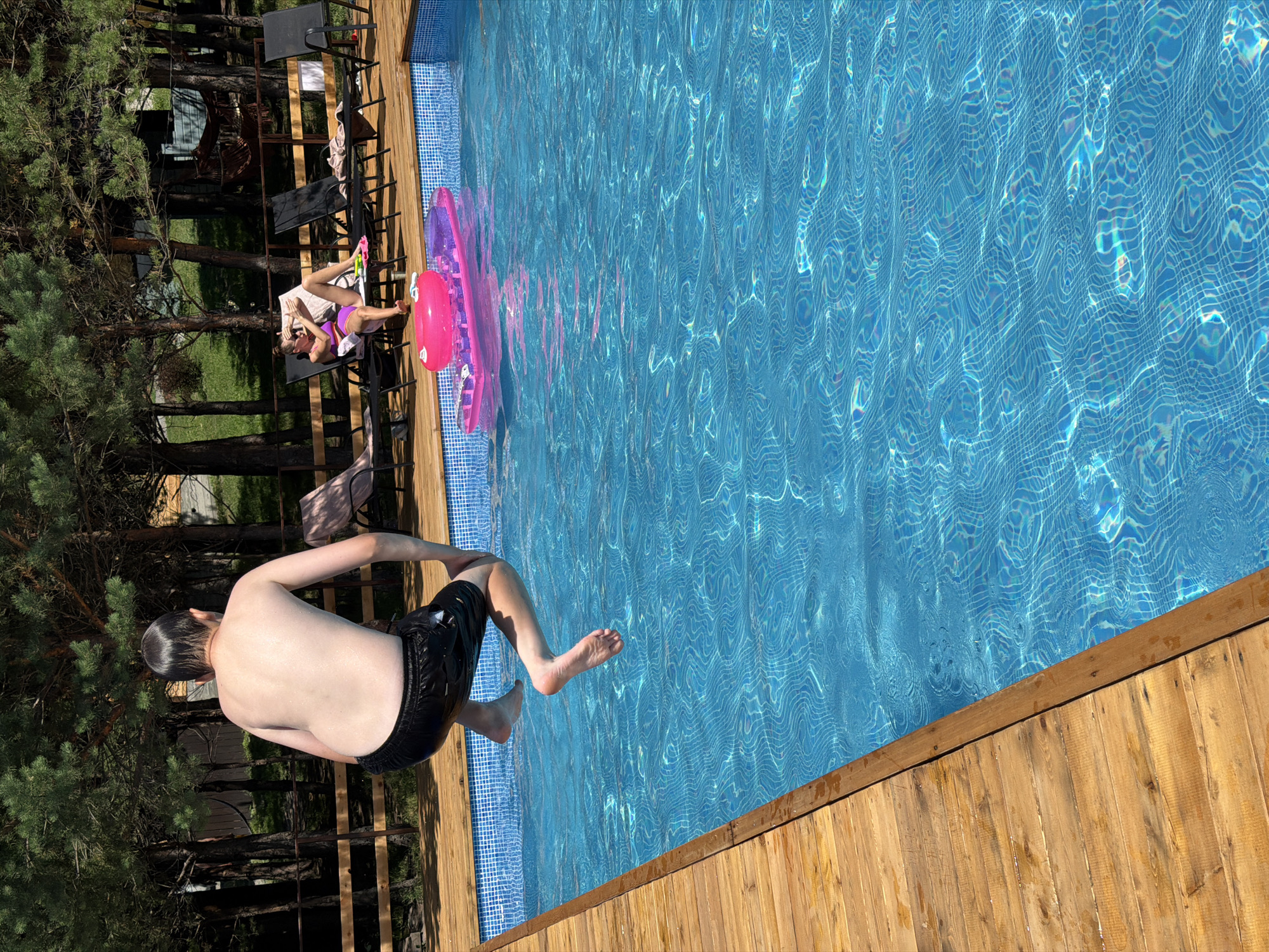 In Elekmonar, Russia, on a bright sunny day, a young boy in black swim shorts is captured mid-air, leaping into a vibrant blue swimming pool. His body is angled downwards, arms extended, and his legs are bent, indicating the momentum of his dive. The pool water, with its characteristic mosaic tile lining visible at the edges, reflects the sunlight, creating a shimmering, dynamic surface. A wooden deck surrounds the pool, adding a rustic, natural element to the scene.
In the background, a woman reclines on a dark sun lounger, dressed in a purple bikini. She appears to be relaxed, possibly checking her phone, enjoying the warm weather. A large, inflatable pink mattress floats in the pool near her, suggesting leisure and fun. The area around the pool is dotted with lush green trees, hinting at a natural, possibly forested, environment. The clear blue sky overhead suggests fair weather, ideal for outdoor activities. The overall mood is one of carefree summer enjoyment and active recreation. No visible text is present in the image.