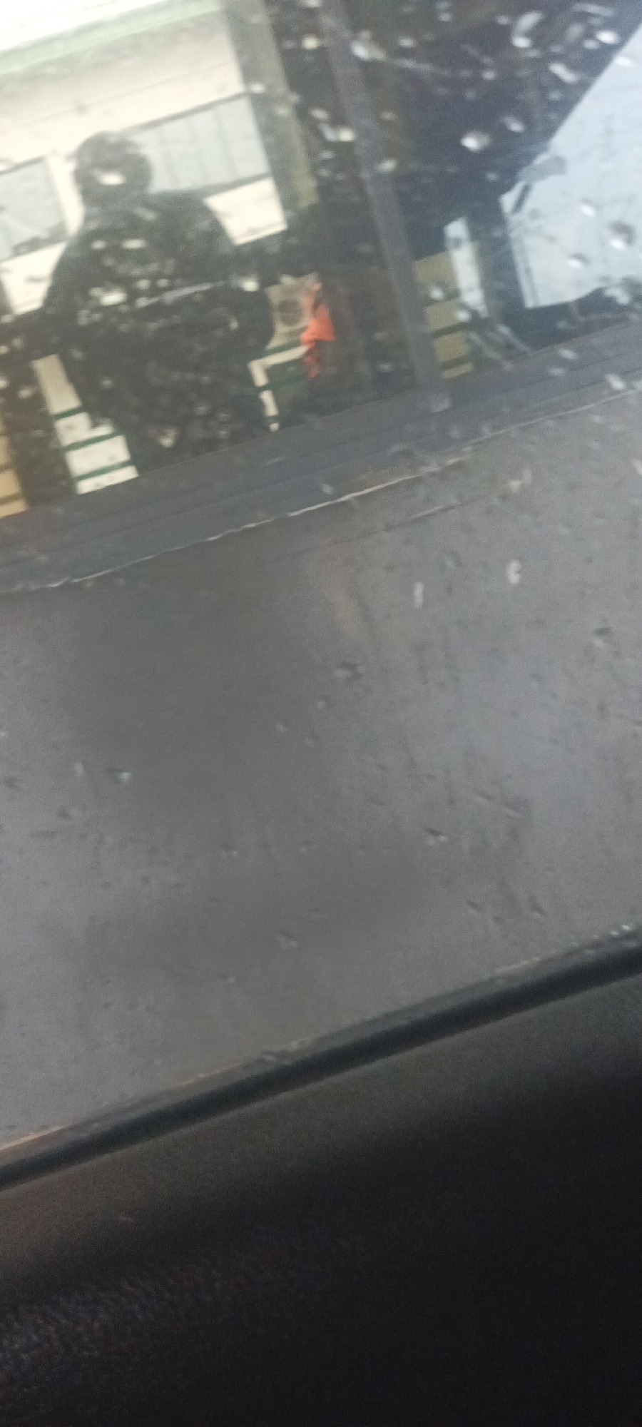 Through a rain-spattered window, a person wearing dark clothing is reflected. An orange object is visible behind them. The location is presumably Kadarko, Nigeria, but there is no visual confirmation.
