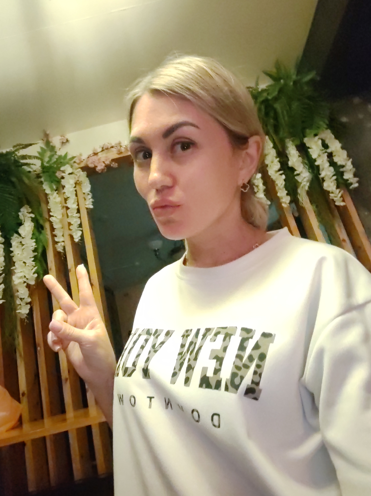 The image depicts a young Caucasian woman with blonde hair, taking a selfie indoors. She is wearing a white sweatshirt with a camouflage-patterned text that appears to read "NEW" in large letters. Her left hand is raised, forming a peace sign. The background shows a wooden structure adorned with artificial white flowers and greenery. The lighting is dim, suggesting an evening or indoor setting. The scene does not contain any specific landmarks or indicators to identify it as being in Komsomolsk-on-Amur, Russia.