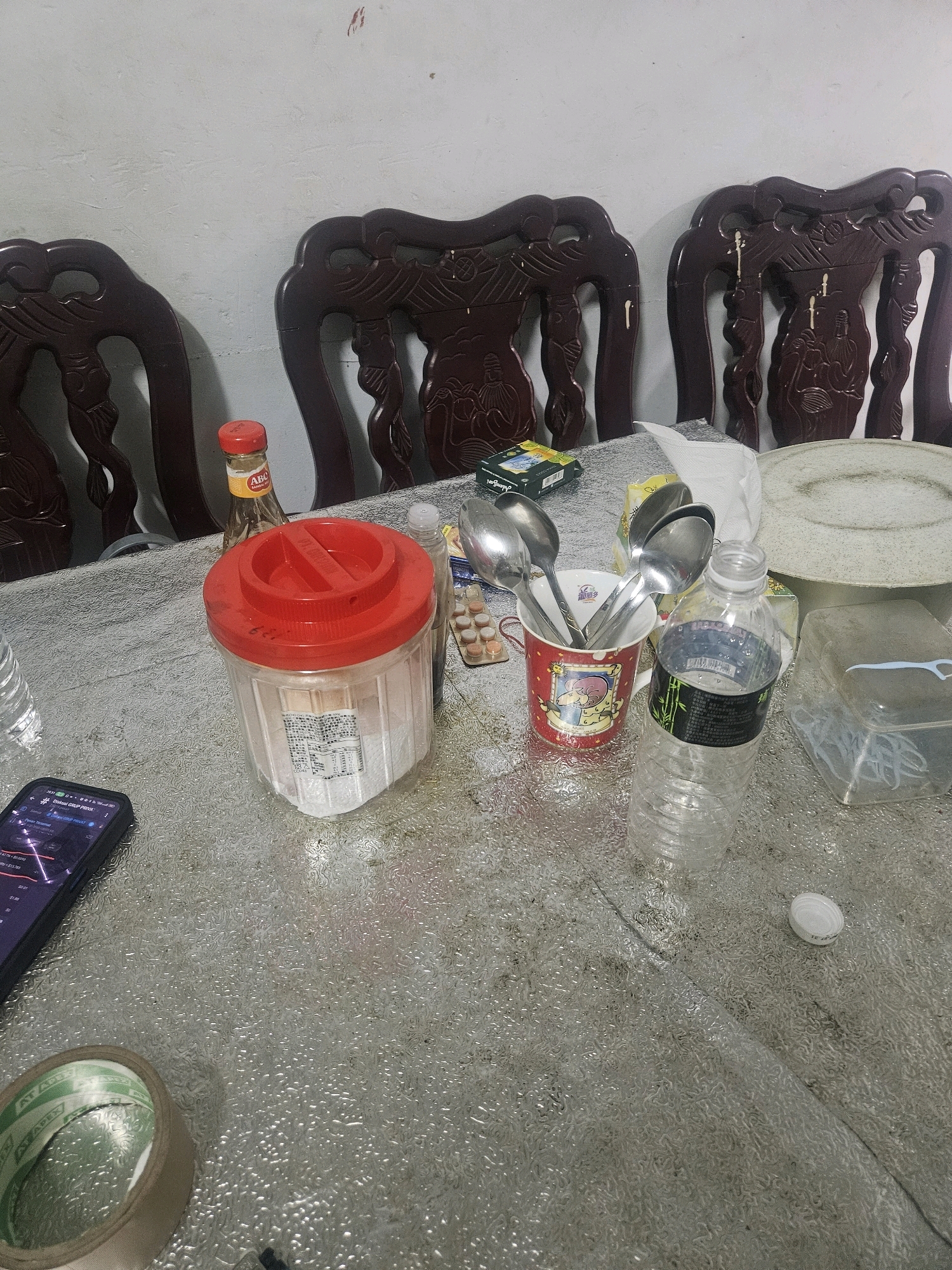 The image shows a close-up, overhead view of a table covered with a textured, silvery tablecloth. On the table, there are several objects, including three ornate, dark wood chairs with carved backs, a large red-lidded plastic container filled with a white powdery substance, a bottle of ABC Sambal, a red mug containing several metal spoons, a plastic water bottle, a small box of medication or supplements, and a clear plastic container with blue dental floss. To the left of the table, a smartphone is visible, displaying a graph on its screen. In the lower left corner, a roll of tape is also present. The background consists of a plain, textured wall with some faint marks. The overall scene appears to be indoors, likely in a residential setting. There are no people visible in the image, and no explicit indication of the location being Huwei Township, Taiwan.