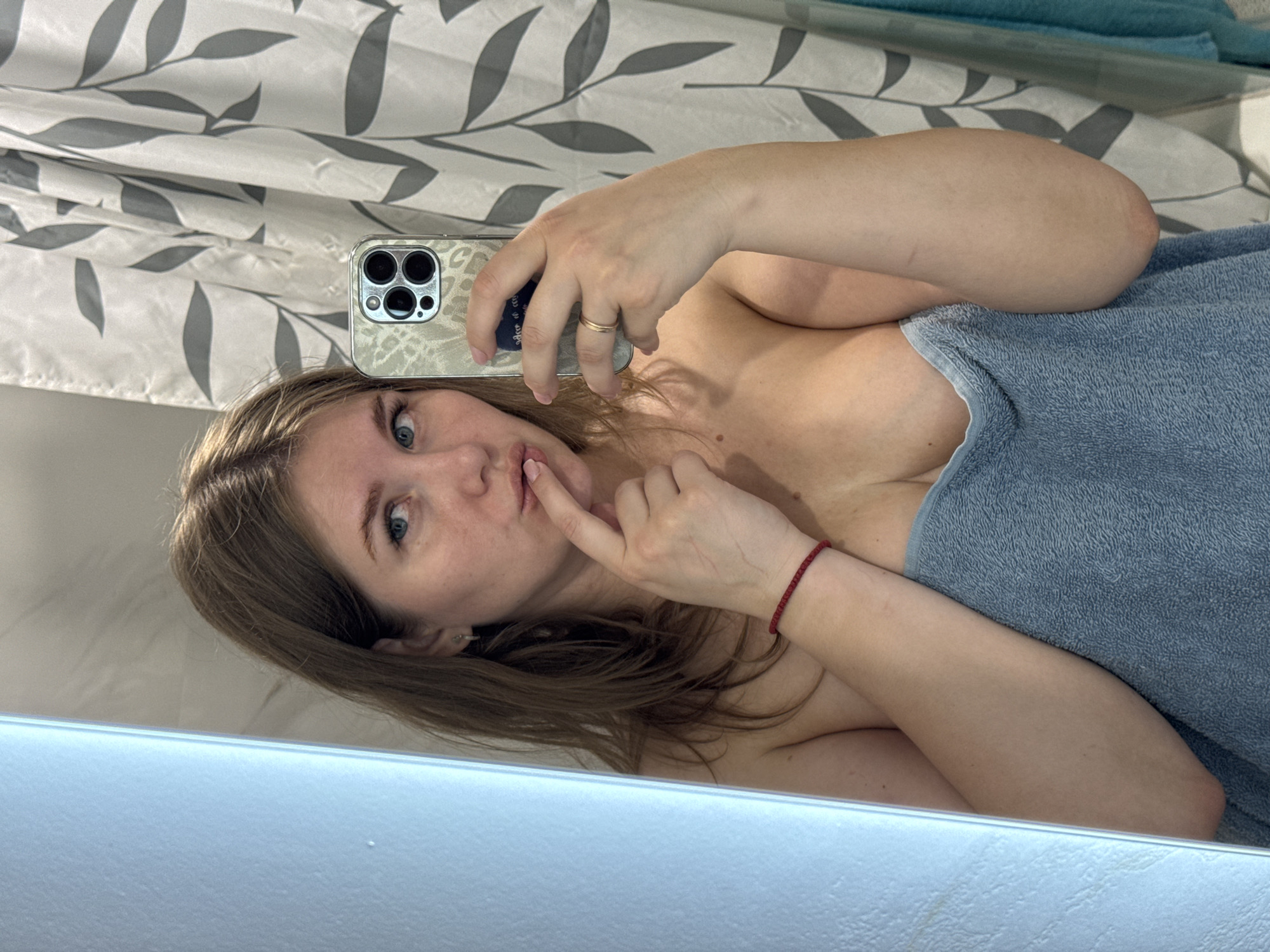 A selfie taken in a bathroom. A woman with fair skin and brown hair holds a smartphone, taking a picture of herself in a mirror. She is partially covered by a blue towel. Her index finger is touching her lips. There is a shower curtain with a leaf pattern behind her. The location is Ферма, Russia.