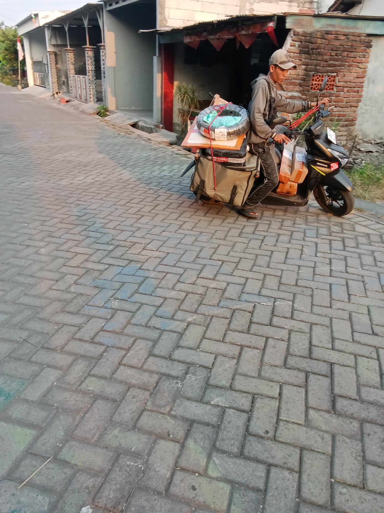 A man wearing a baseball cap, grey hoodie, and patterned pants rides a black scooter down a paved street in Sidorejo, Indonesia. The scooter is loaded with packages and a coiled item wrapped in plastic. The man is looking forward, appearing to be driving. He is on the right side of the road, with buildings and a brick wall visible on the right side of the frame, and more buildings on the left. The street is paved with interlocking bricks. The lighting suggests it is either early morning or late afternoon.