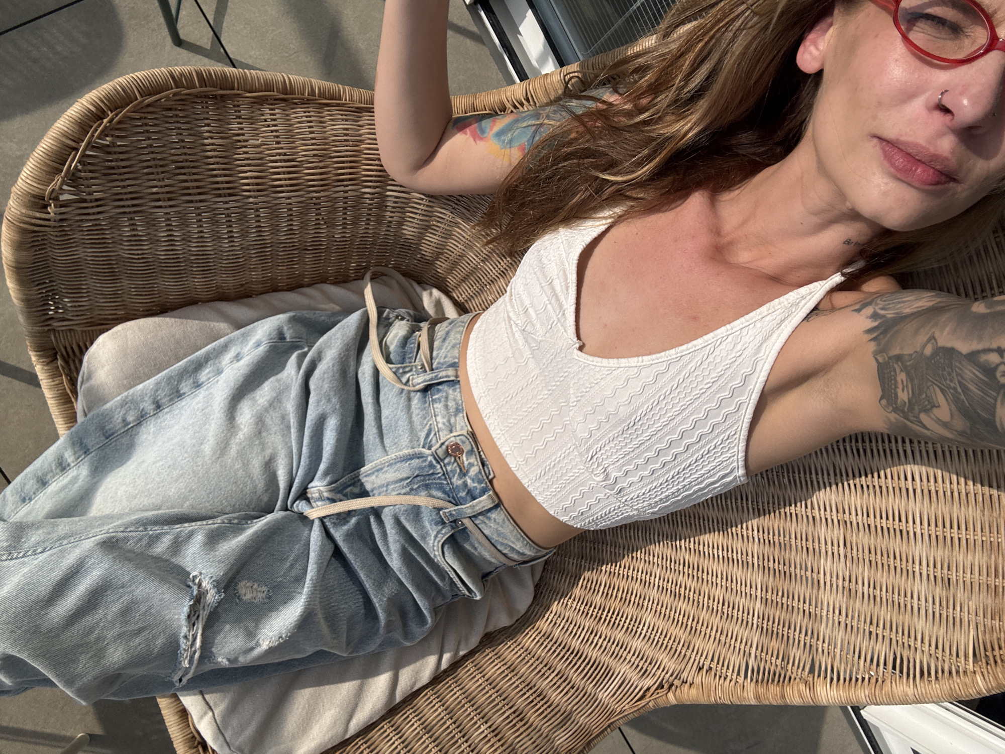 A woman with fair skin, wearing red glasses and a nose ring, is reclining in a woven wicker chair outdoors. She has light brown hair and is wearing a white, textured crop top and light blue, high-waisted jeans with a drawstring. A tattoo is visible on her left arm and another on her right arm near her shoulder. The chair is situated on a flat surface, likely a balcony or patio, with sunlight casting shadows. The location is presumed to be Tel-Aviv, Israel.
